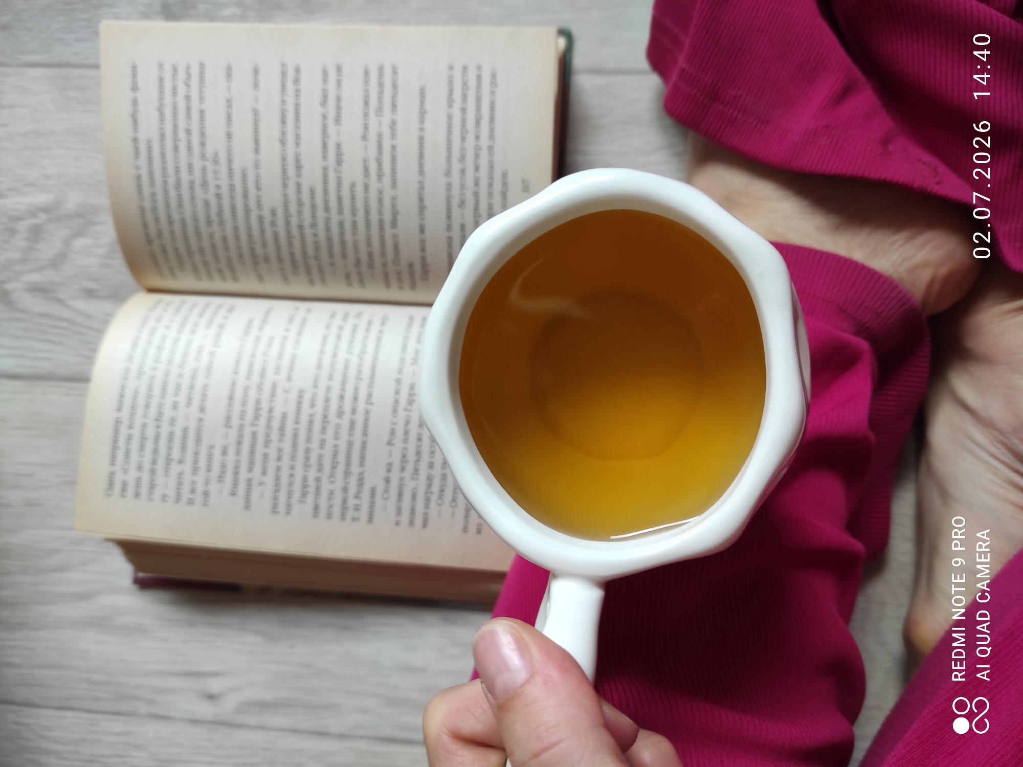 A first-person perspective shot captures a white mug filled with amber liquid, likely tea, held by a hand with visible fingernails. The mug rests on a surface of bright pink fabric. In the background, an open book with text is visible, its pages are slightly worn. The environment appears to be an indoor setting with a wooden floor visible beneath the fabric. A timestamp in the lower right corner reads "02.07.2026 14:40", and a watermark in the lower left indicates "REDMI NOTE 9 PRO AI QUAD CAMERA". There is no identifiable information or context related to the city of Malyi Bukor, Russia in the image.