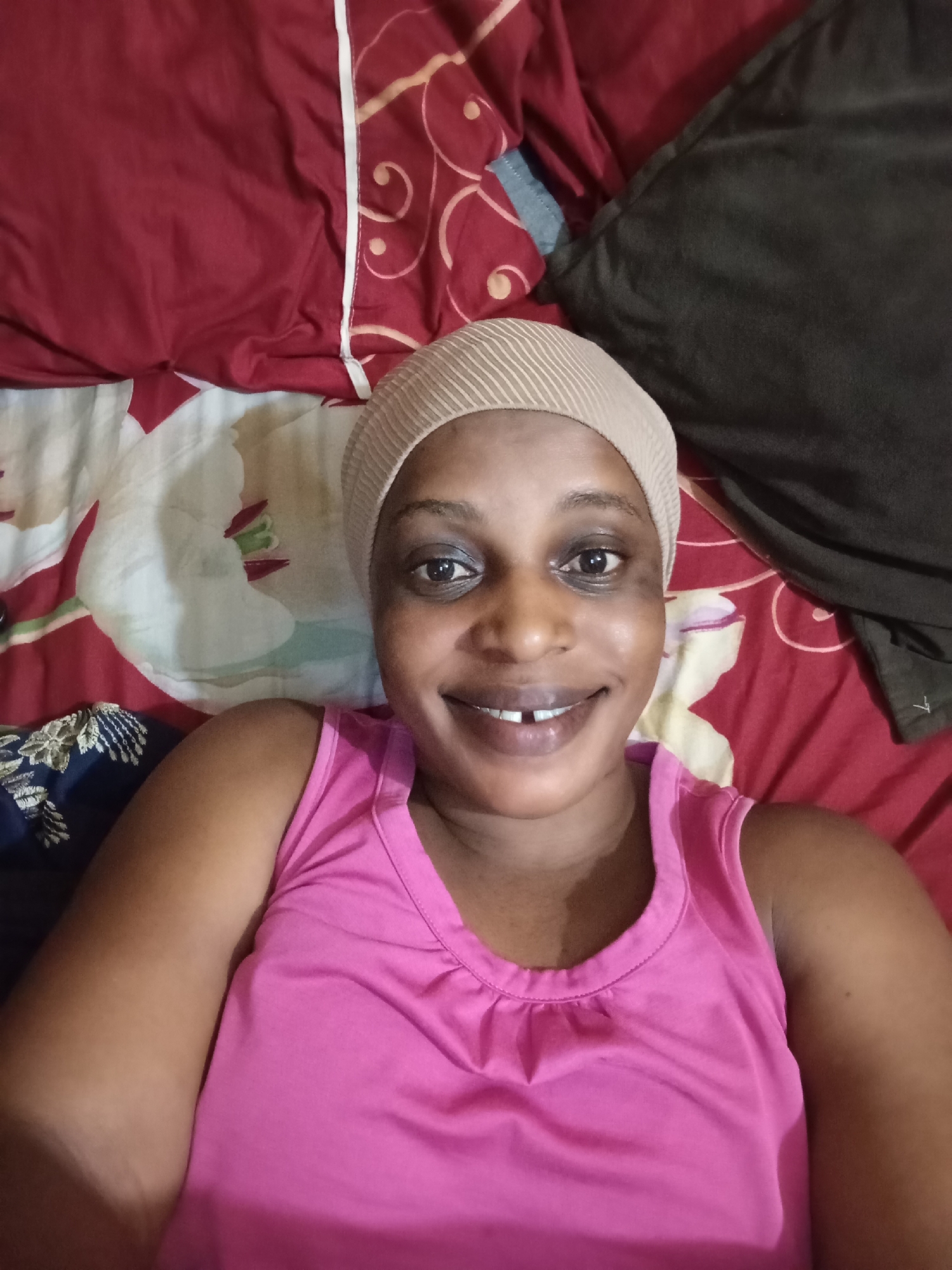 A medium shot shows a woman with dark skin, wearing a pink tank top and a beige striped head covering. She is smiling and lying on a bed. The bedding has a busy pattern of reds, whites, and browns. To the left of the frame is a small section of blue and gold fabric, and to the right, a dark gray pillow. The image was taken in Narayi, Nigeria.