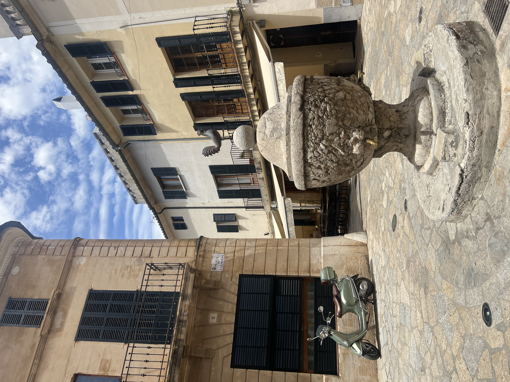In Pollença, Spain, an outdoor square features a stone fountain with a rooster weather vane. A light green scooter is parked to the left of the fountain. Buildings with closed shutters flank the square, and a street sign for "PLACA DE L'ALMOINA" is visible on the corner of one building. The ground is paved with irregular stone tiles. A few tables and chairs are set up at an outdoor cafe in the background, and a person is seated at one of the tables. The sky is partly cloudy.