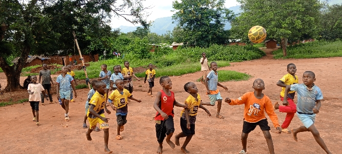 The scene depicts approximately eighteen children, predominantly young boys appearing to be between 5 and 12 years of age, engaged in playing football on a reddish-brown dirt field in Ngalula, Democratic Republic of the Congo. A yellow and black patterned football is airborne in the upper-middle right quadrant of the image.
The children are dressed in various casual attire including t-shirts, shorts, and pants; several wear shirts with sports logos such as "1&1" and "BVB" (Borussia Dortmund), and "Vodafone". Many of the children are barefoot, while some wear simple shoes or sandals. Their expressions indicate active engagement in the game, with several looking up towards the ball or towards other players.
In the foreground, a child in an orange long-sleeved shirt and black shorts looks directly upwards towards the ball, with his arms slightly raised. To his right, a child in a light blue t-shirt and shorts is captured mid-stride, running with his gaze also directed towards the ball. Another child in a red sleeveless top and black pants runs towards the right.
The environment includes a dense backdrop of green foliage, large trees, and bushes, with distant mountains visible on the horizon under a light sky. Several rudimentary structures, appearing as small huts or houses with simple roofs, are scattered within the background vegetation. A tall, slender bamboo-like pole stands upright on the left side of the frame. The overall setting suggests a rural or semi-rural community.
