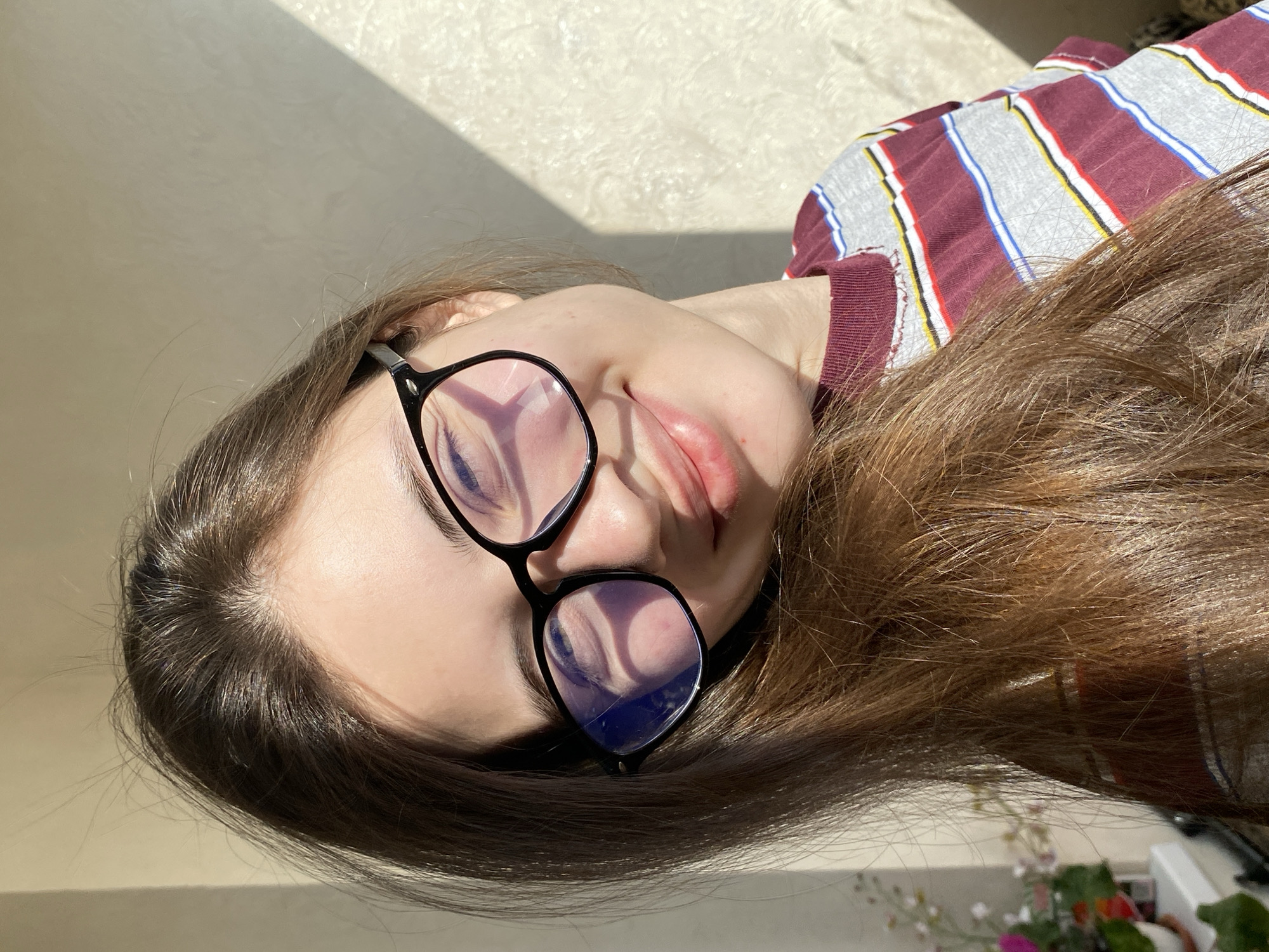 A young woman is captured in a close-up, lying on her side with her head slightly tilted towards the viewer. Her long, rich brown hair is spread out, partially obscuring her ear and shoulders. She wears black-rimmed glasses, and her eyes appear closed or deeply relaxed, with her lips softly parted. Strong, directional sunlight illuminates her features and hair, creating sharp shadows. Geometric light patterns, likely reflections from a window, are prominently visible on the lenses of her glasses. She is dressed in a multi-striped top featuring maroon, grey, white, blue, and yellow bands. The background behind her consists of a plain, light-colored wall, across which a distinct diagonal shadow falls. In the lower right corner, a potted plant with colorful blooms and blurred household objects are visible, indicating an indoor domestic setting. The scene suggests a moment of rest or sleep on a sunny day in Votkinsk, Russia. No discernible text is present.