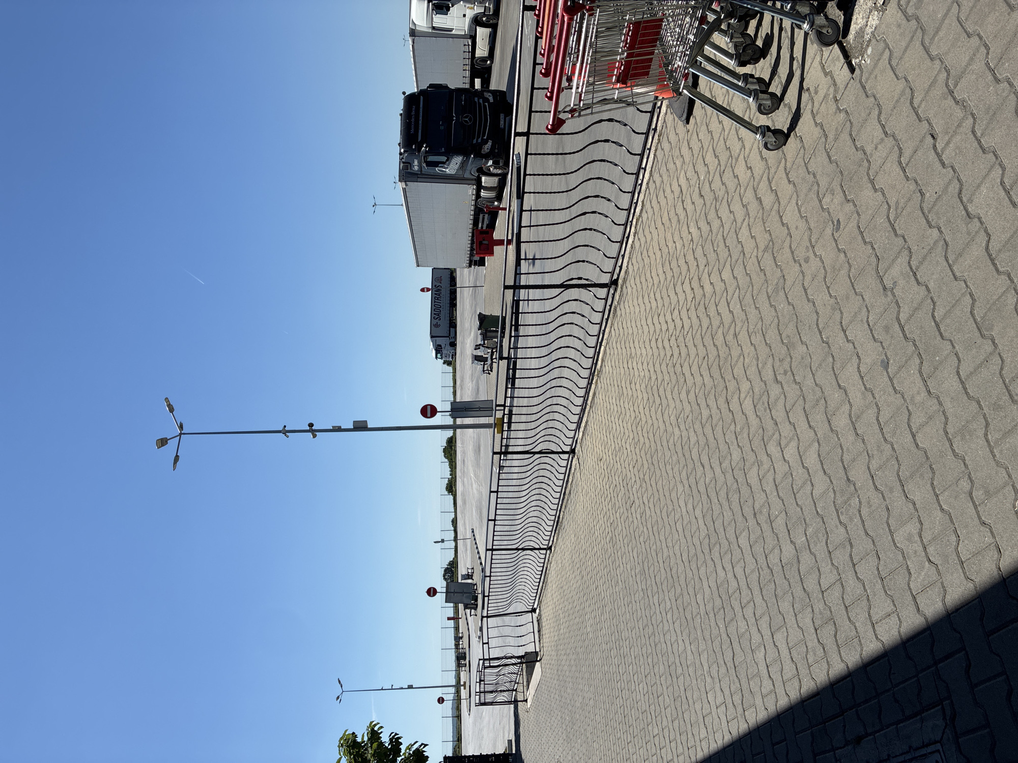 The image shows a sunny outdoor scene, possibly in a parking or loading area, located near Lesovo, Bulgaria. The ground is paved with interlocking concrete blocks. A black, decorative, wave-patterned metal fence separates the paved area from a loading zone containing several large trucks, including a black Mercedes-Benz semi-trailer. To the right of the fence, a row of shopping carts is visible. Light poles with three directional lamps extend upwards towards a clear, blue sky. A thin contrail cuts through the sky in the distance. In the background beyond the trucks, other parking or storage areas are visible, delineated by a wire fence. Stop signs on metal poles are present.