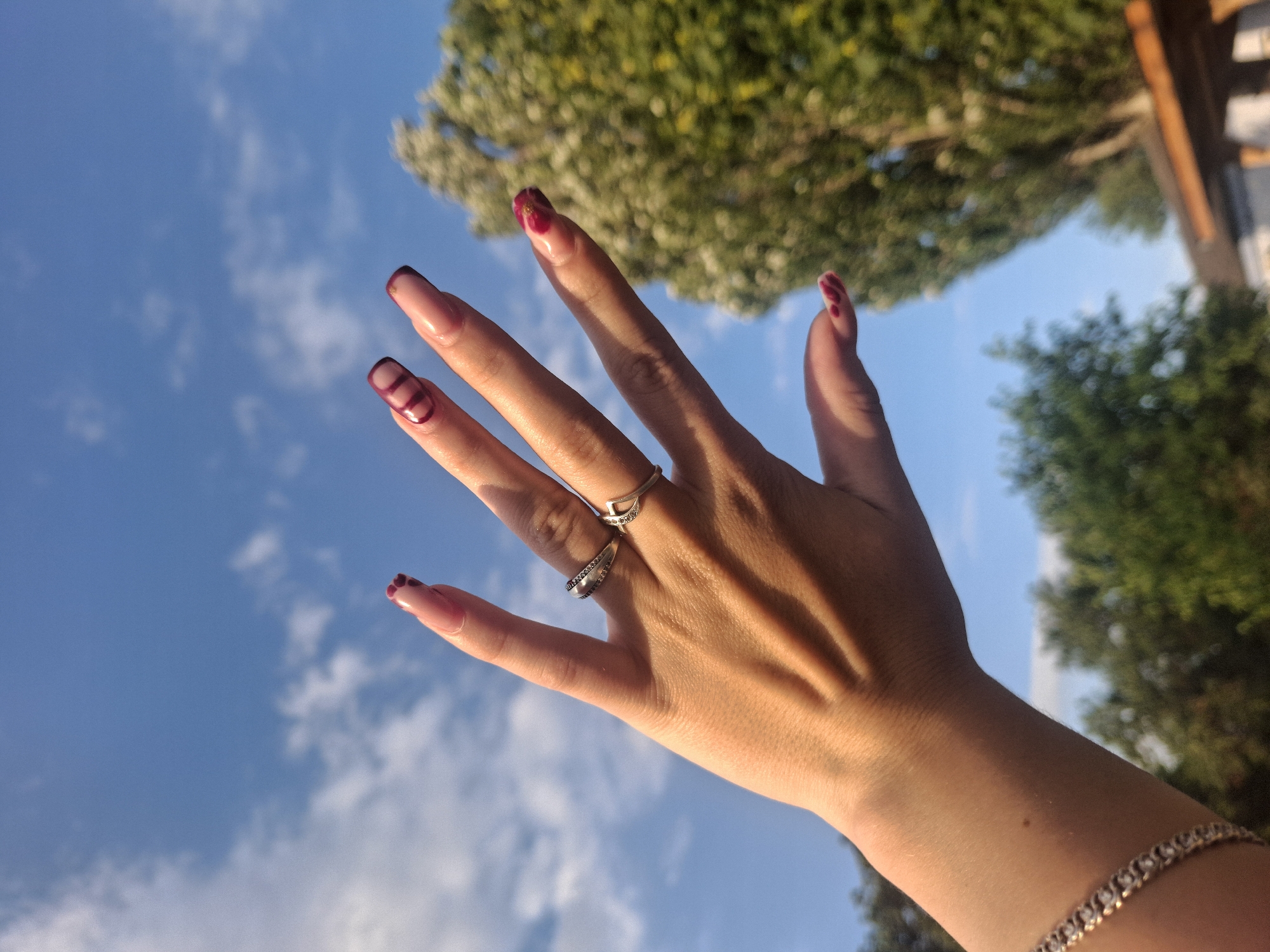 A woman's hand with long, manicured nails, adorned with burgundy-colored polish and decorative lines, is held up against a bright blue sky with scattered white clouds. The hand wears two silver rings, one with small, dark stones and the other shaped like a crescent moon. A silver bracelet is visible on the wrist. In the background, lush green trees are blurred, and a portion of a wooden structure is visible at the bottom right. The lighting suggests it is daytime.