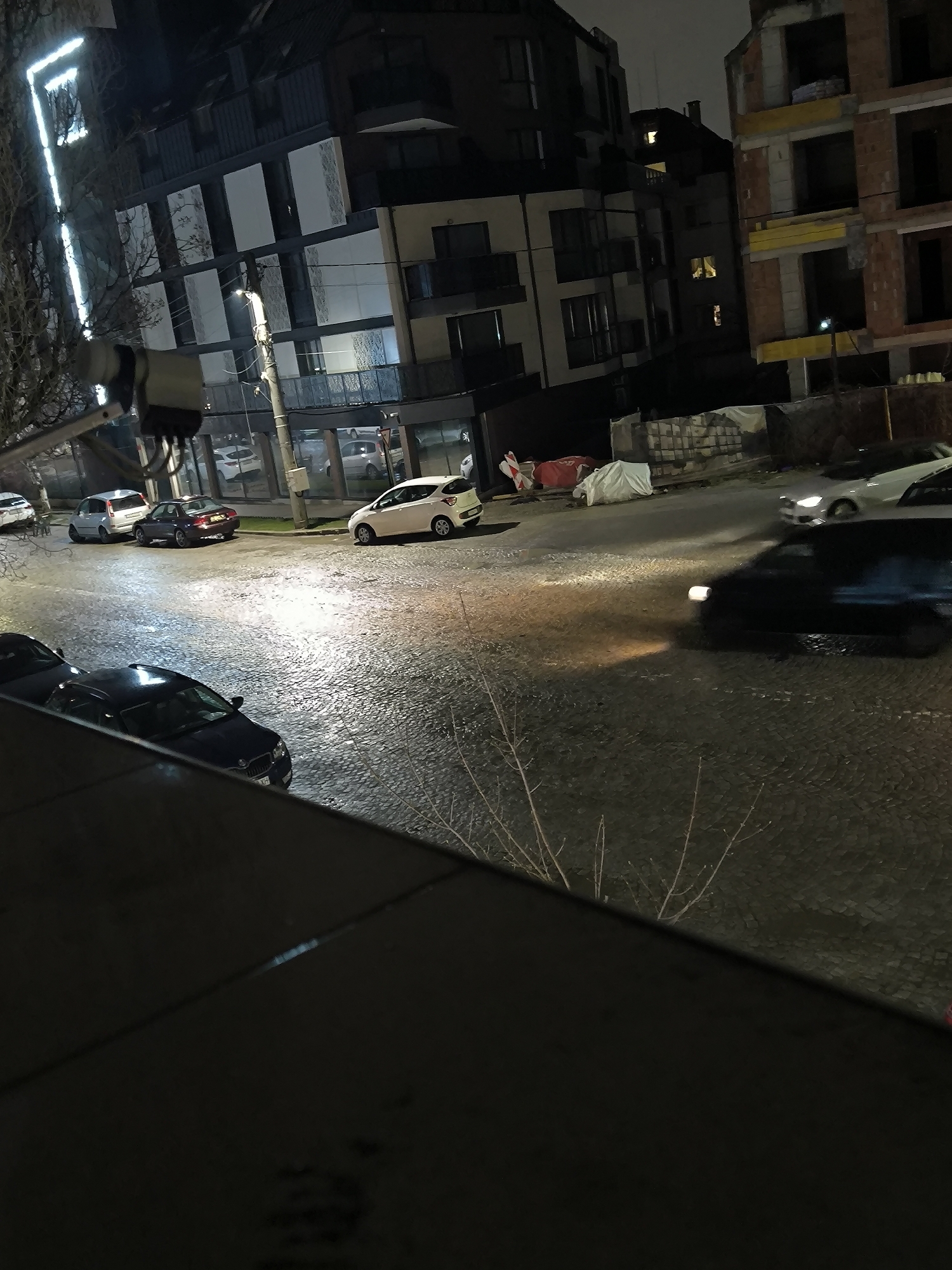 The image shows a street scene in Sofia, Bulgaria, at night. The street is paved with cobblestones and appears wet, likely due to rain. Several cars are parked or driving on the street. Buildings line the street, including a modern building with illuminated architectural elements and an unfinished brick building under construction. Construction materials covered with white tarps can be seen near the incomplete building. Branches from nearby trees are visible in the foreground.