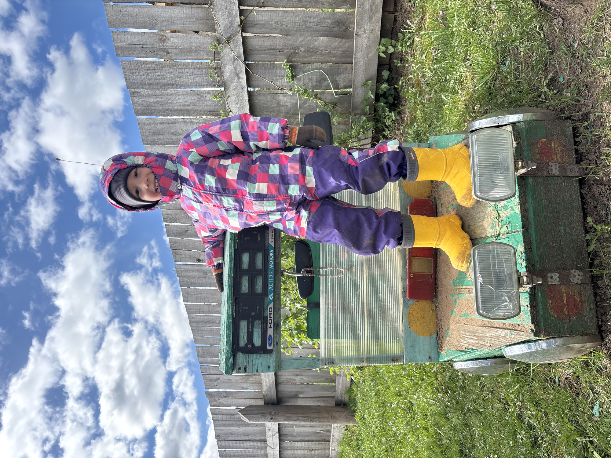 A child in a multicolored snowsuit and yellow boots is positioned on a structure, likely a homemade toy vehicle or playhouse, outdoors. The structure is painted in green and blue hues, with attached features resembling headlights and a license plate frame that reads "Ford." The background includes a wooden fence and green foliage, suggesting a backyard or garden setting. In the sky are white clouds. The location is the city of Ваньки, Russia.