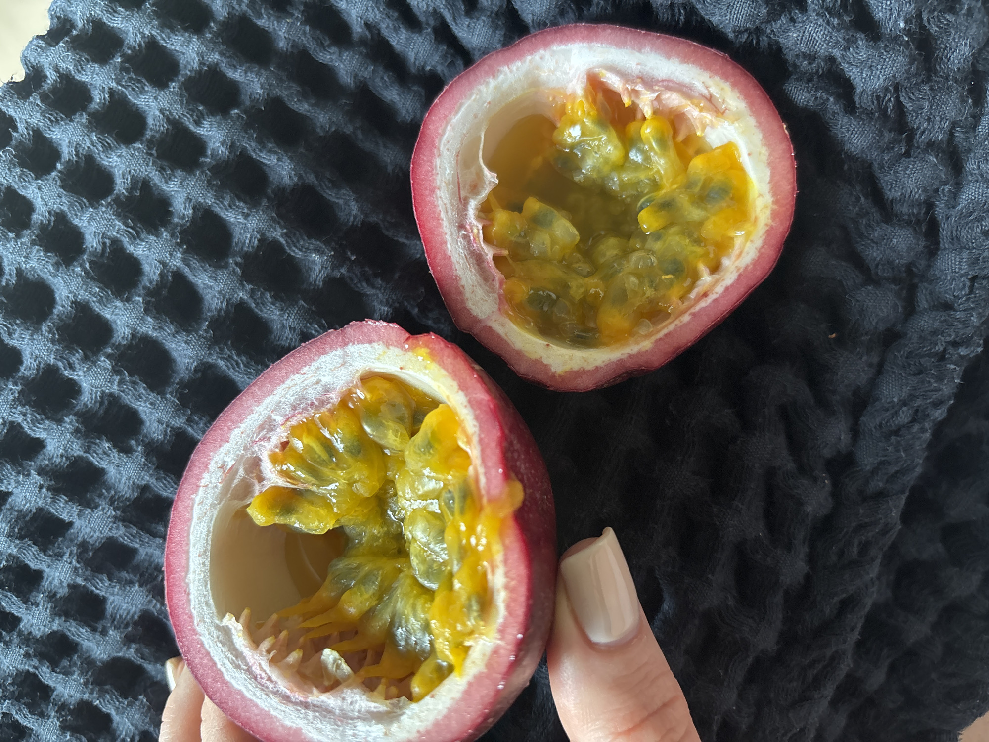 Two halves of a passion fruit are displayed on a black, textured cloth. The fruit halves show the interior yellow pulp with dark seeds. A hand with a light-colored fingernail is holding the bottom half of the fruit.