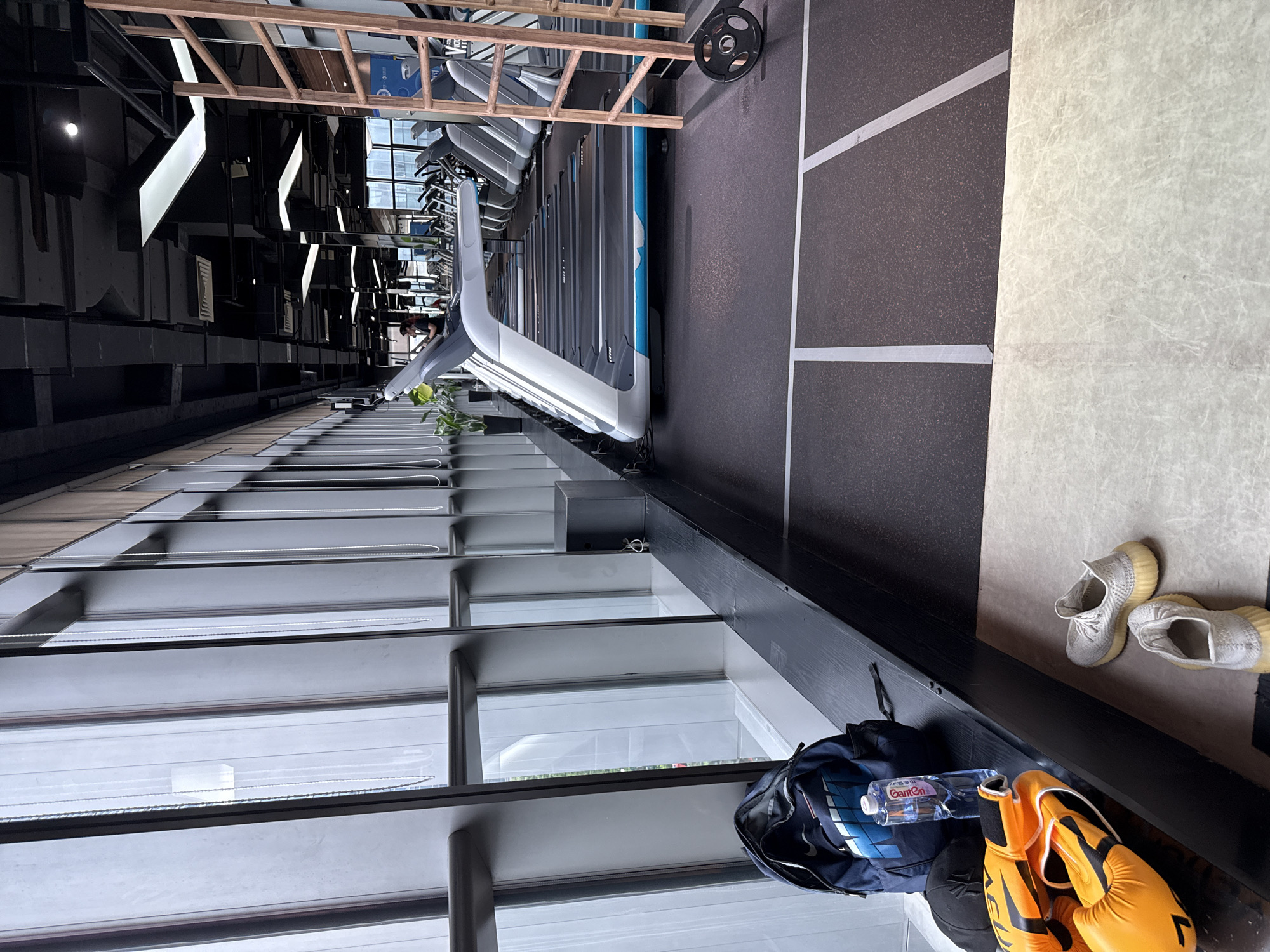 This image depicts the interior of a modern gym, likely located in Haizhu District, China, with large windows providing a view of an urban setting. In the foreground, a navy blue backpack with a blue and white Nike logo rests on a dark, wooden bench. Next to the backpack are an orange pair of boxing gloves and a black baseball cap. A clear plastic bottle of water is also placed on the bench. Further to the right on the floor, a pair of white athletic shoes are visible. The mid-ground of the image is dominated by a row of treadmills, with their grey and blue frames extending into the distance. A woman is faintly visible operating one of the treadmills. To the right of the treadmills, a tall, wooden ladder-like structure stands near a gym weight. The overall environment is clean and organized, with black ceilings, exposed ductwork, and bright, recessed lighting contributing to the contemporary aesthetic of the gym.