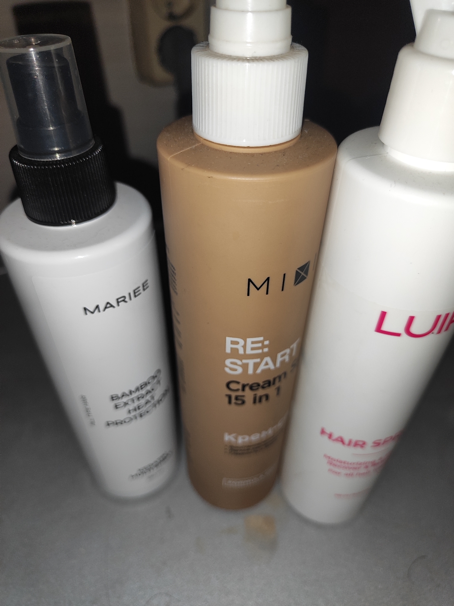 Three bottles of hair products are visible in the foreground. The bottle on the left is white with a black pump dispenser and a clear cap. Text on the bottle reads "MARIEE BAMBOO EXTRA HEAT PROTECTOR". The middle bottle is light brown with a white pump dispenser. Text on this bottle reads "MI RE: START Cream 15 in 1 Kpemp". The bottle on the right is white with a white pump dispenser. Pink text on this bottle reads "LUI HAIR SP". The bottles are arranged diagonally from left to right and are the primary focus of the image. The background is dark and out of focus, showing what appears to be a wall outlet. The lighting is dim.