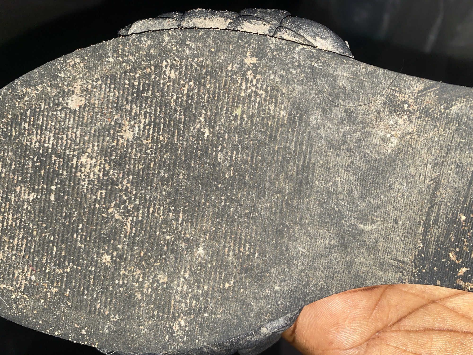 The image shows the sole of a dirty shoe, possibly a sandal, held by a person's hand. The sole is made of dark rubber with horizontal tread marks. It is covered in a light-colored dust or dirt, with small particles embedded in the grooves. The edge of the sole shows a woven or braided texture, suggesting it is part of a sandal. The hand holding the shoe is partially visible on the left side of the image, with clear fingerprint lines. The background is dark and out of focus, providing no contextual information about the location. The scene is consistent with an everyday outdoor environment where shoes can become dirty.