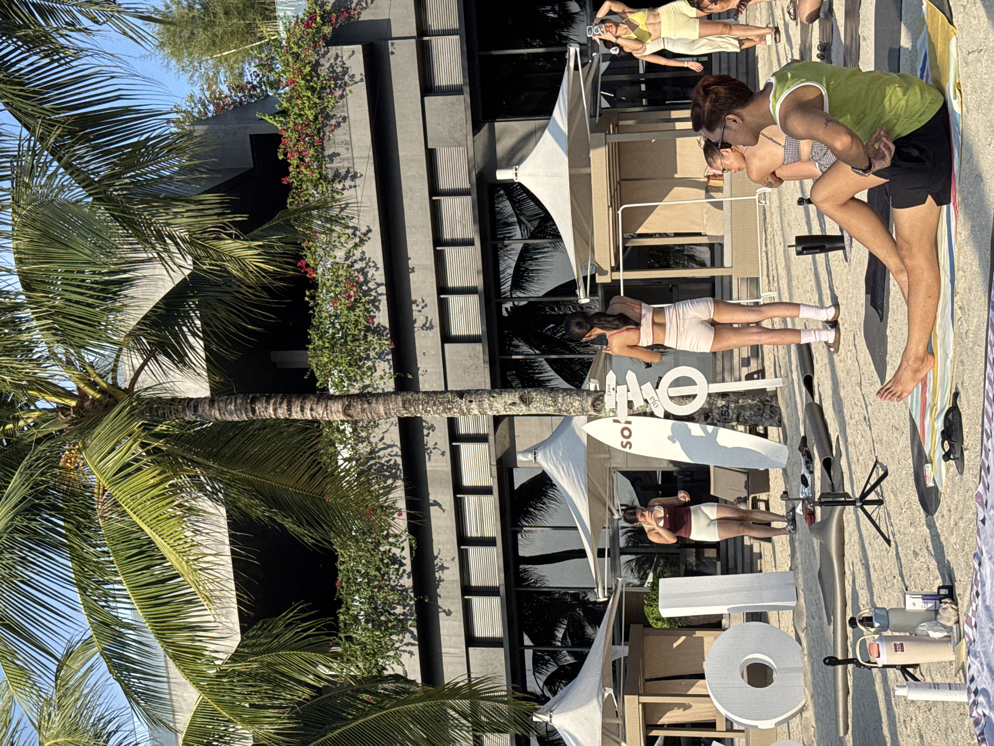 A group of people are gathered on a sandy beach in front of a modern building with large glass windows and balconies adorned with greenery. Palm trees with lush fronds sway in the background. Some individuals are dressed in athletic wear, suggesting a fitness or yoga activity.
In the foreground, a man wearing a green tank top and black shorts is seated on a striped mat, facing away from the camera and towards the building. He appears to be engaged in a stretching or meditation pose. To his left, several water bottles, sunscreens, and other personal items are arranged on the sand. A tripod with a small camera is also visible.
Further back, two women are standing near a white surfboard with the word "SOL" visible. One woman, wearing a white sports bra and beige shorts, is holding a drink. Another woman, in a yellow top and white shorts, is holding up her phone, possibly taking a photo. A person in a white tank top and white shorts is also present, standing and looking at her phone. Behind them, other individuals are seated or standing, some with their backs to the camera.
The overall scene suggests an outdoor gathering, possibly a yoga session or a promotional event, taking place on a beach in Samal, Philippines, given the tropical vegetation and beach setting. The lighting indicates it is daytime, with clear skies.