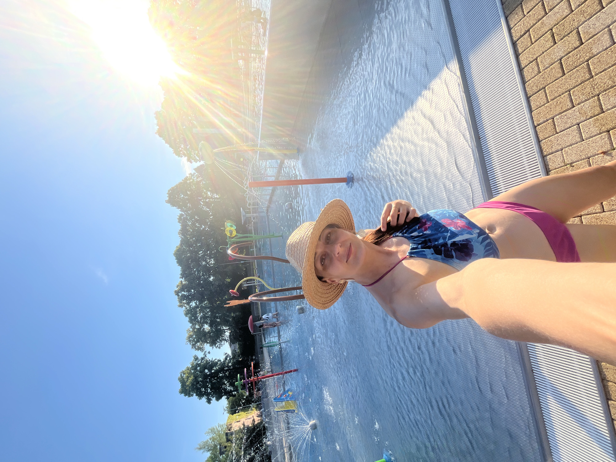 A woman takes a selfie at a splash pad. She is wearing a straw hat, a blue and pink patterned bikini top, and pink bikini bottoms. The splash pad is filled with shallow water and features several colorful spray features. Trees and a bright sun are visible in the background. The location is the city of Unknown, Unknown.