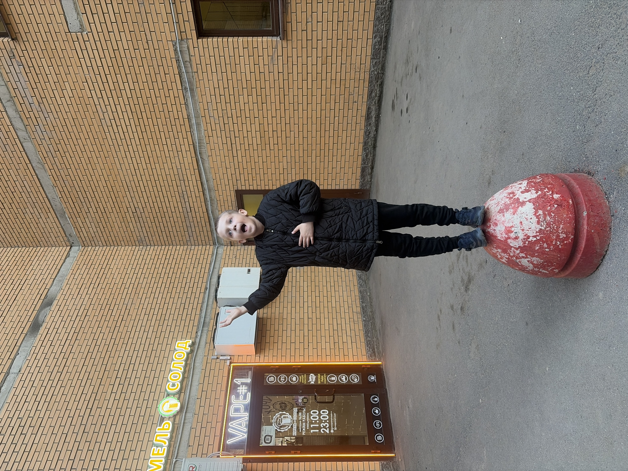 A child stands on a red and white-painted concrete bollard outside a store called "VAPE #1", possibly in Saint Petersburg, Russia. The store has a neon-lit sign and a clear door through which interior details are visible. The store's operating hours, as displayed on the door, are from 11:00 to 23:00. The store is named "Khmel Solod" (ХМЕЛЬ СОЛОД) and the main store sign says "MEL SODOD" (МЕЛЬ СОЛОД). The exterior of the building is brick, and there are air conditioning units mounted on the wall above the storefront. The surrounding ground is paved. The child is wearing a black jacket and black pants and looks animated with an open mouth and outstretched arm.