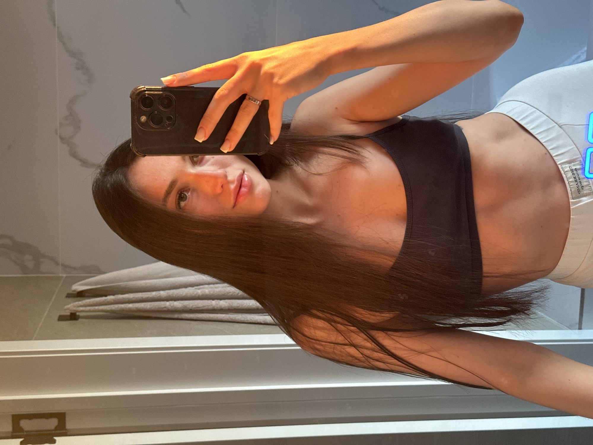 A young woman with long, dark hair takes a selfie in a bathroom. She is wearing a dark sports bra and light-colored pants. Her left hand is holding her phone, and her right hand is resting on the phone, showing a ring on her ring finger. The background includes a tiled wall, towels hanging on hooks, and a shower door. The lighting suggests it is either sunrise or sunset, casting a warm glow on her face and arm. The image appears to be taken in a residential bathroom.