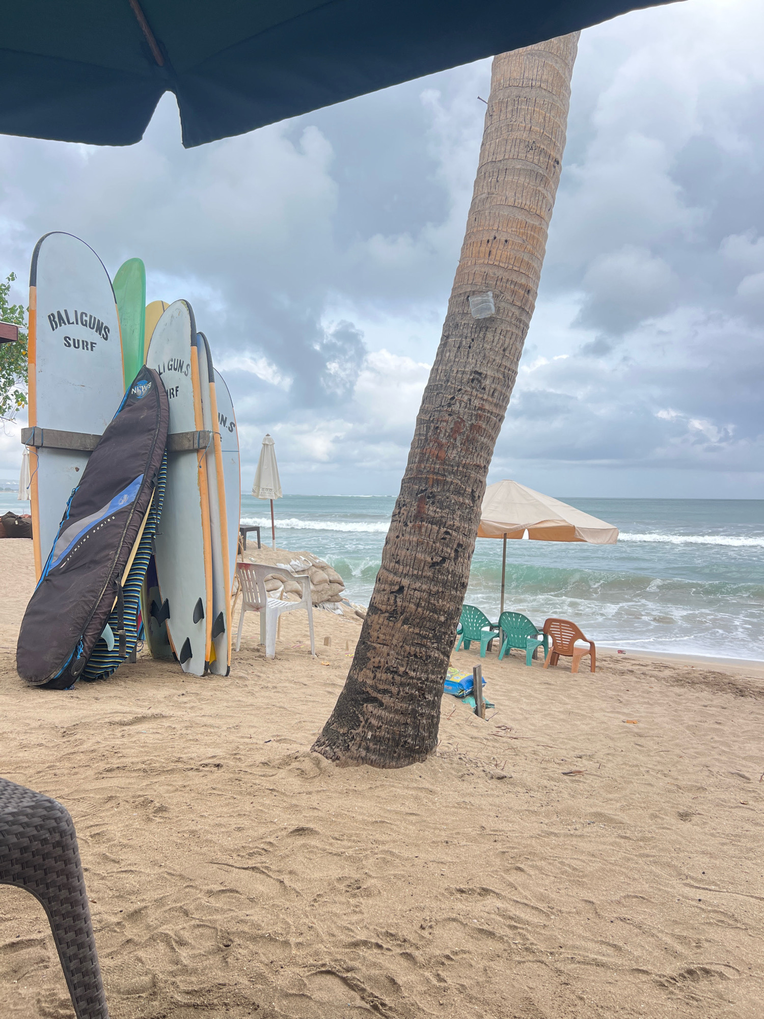 The image captures a beach scene in Kuta, Indonesia, under an overcast sky. In the foreground, a dark wicker chair is partially visible on the sandy beach. To the left, a collection of surfboards stands upright, leaning against a wooden rack. One surfboard is enclosed in a dark protective bag. A tall, textured palm tree trunk dominates the right side of the frame, obscuring part of the ocean view. Beyond the palm tree, beach chairs and a large umbrella are set up on the sand near the water's edge. The ocean waves are actively breaking on the shore, and the water appears greenish-blue. In the background, the horizon is marked by a cloudy sky.