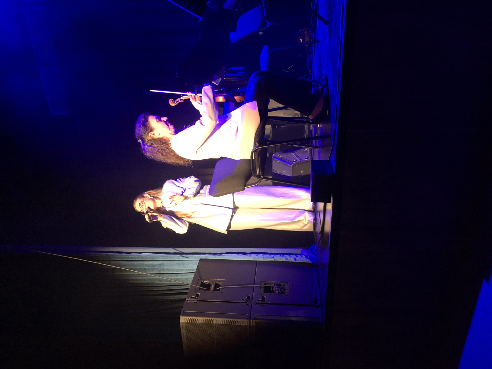 A woman sings into a microphone on stage, wearing a white suit and glasses. To her right, another woman sits and plays the violin. The stage is lit with blue and purple lights, and a dark curtain hangs behind the performers. Two large speakers are visible on the left side of the stage. The background is dark, suggesting an indoor performance space, possibly a concert hall or theater. This scene appears to be from a performance at the Moscow Conservatory in Moscow, Russia.