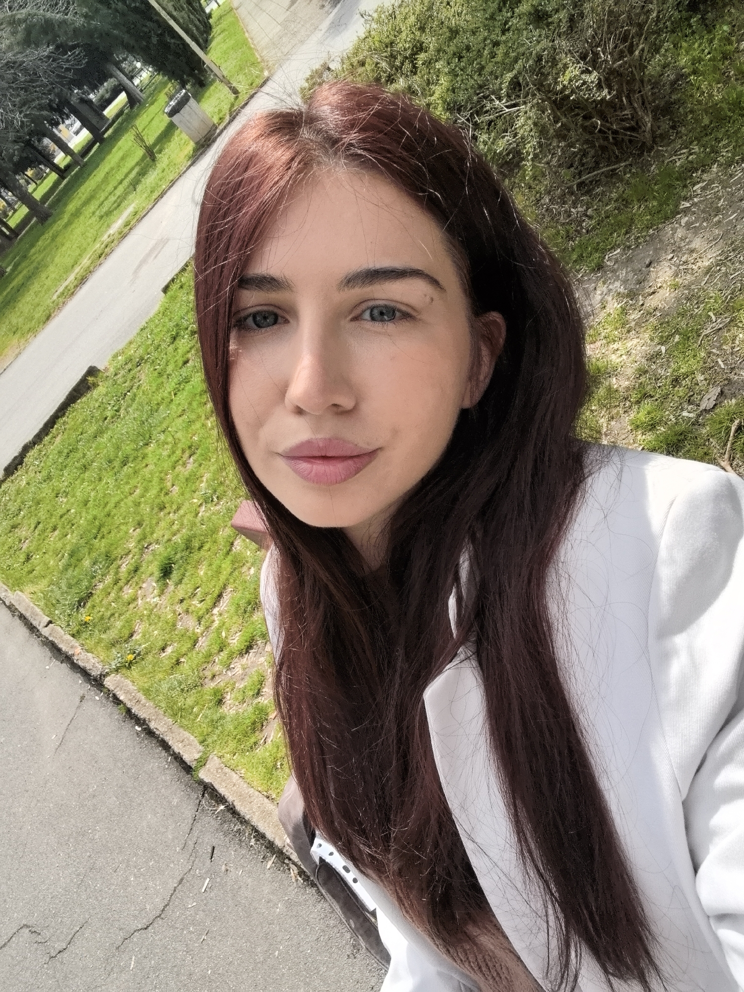 The image is a selfie of a young woman with long, auburn hair. She has fair skin, blue eyes, and pink lips. She is wearing a white blazer. The background shows a park-like setting with green grass, trees, and a paved walkway. A trash bin is visible in the background. Based on the visual context of the image, it is likely that the location is a park or garden within the city of Stara Zagora, Bulgaria.