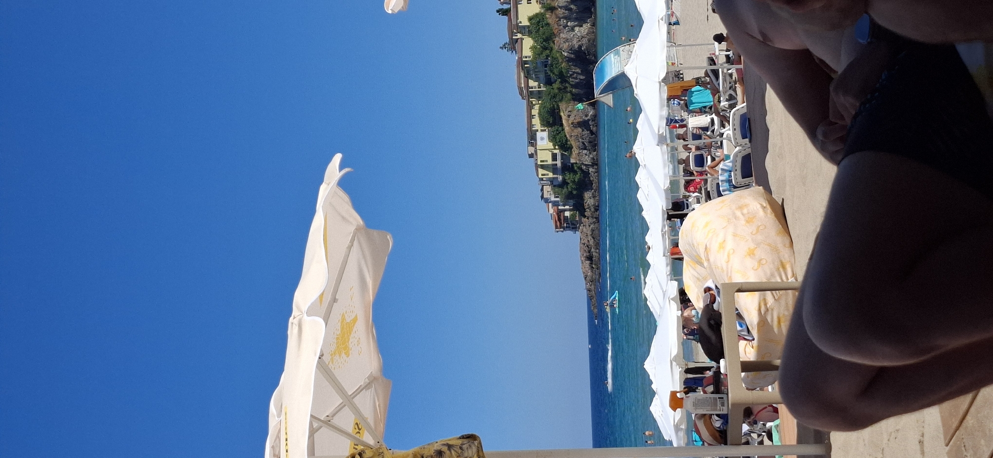 A sun-drenched beach scene unfolds in Sozopol, Bulgaria, under a clear blue sky. The foreground is dominated by the legs of a person lounging on the sand, partially obscured by a large, patterned beanbag chair. To the left, a table holds various beach essentials, including a bottle of sunscreen and a dark object, possibly a towel or clothing. Beyond this, rows of white beach umbrellas stretch across the sandy expanse, shading numerous lounge chairs where beachgoers relax. The middle ground features the turquoise waters of the sea, dotted with swimmers and a few watercraft. In the distance, a rocky coastline is topped with buildings, their yellow facades contrasting with the lush green vegetation. A white and blue structure, likely a part of a beach attraction, stands near the shore. The overall atmosphere is one of a tranquil summer day at a popular seaside resort.