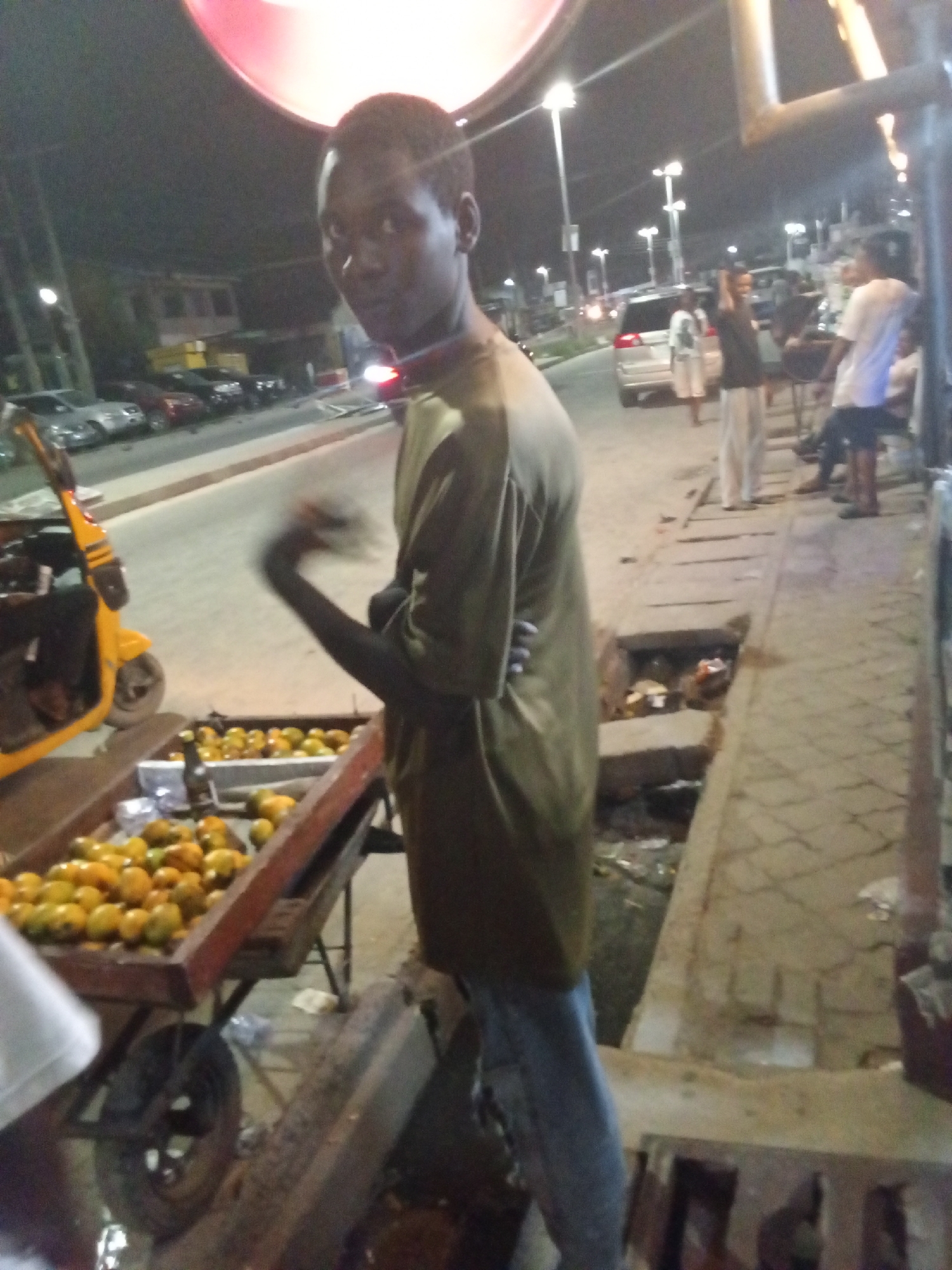 This image depicts a bustling street scene at night in Ikorodu, Nigeria. In the foreground, a young man, likely a vendor, stands next to a wooden cart filled with numerous oranges or similar citrus fruits. He wears a dark, long-sleeved shirt and blue jeans, looking towards the camera with a neutral expression; his right arm is slightly blurred, suggesting motion.
To his left, a bright yellow tricycle (Keke Napep) is parked. The street behind him is illuminated by multiple bright streetlights, revealing several cars parked or in motion. Various other people are visible in the mid-ground and background, gathered on the street and sidewalk, contributing to the lively evening atmosphere. The paved sidewalk runs alongside an open drainage channel, which contains some debris. A large, circular red-lit sign is partially visible at the top of the frame. The overall image has a somewhat blurry and grainy appearance, consistent with low-light photography. No specific text is clearly legible. The scene captures a common urban street-side commerce and social activity after dark.