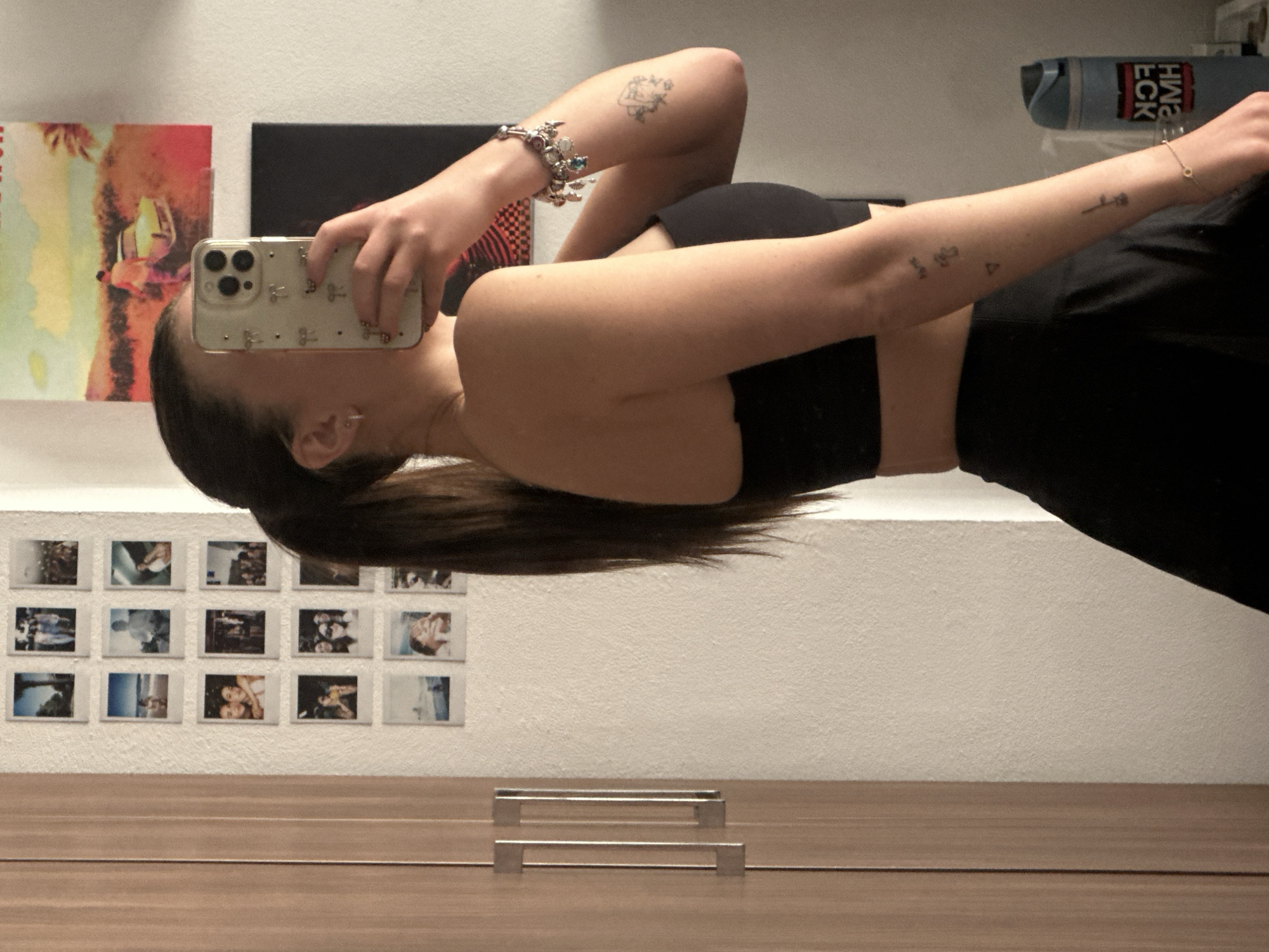 A woman is taking a selfie in a mirror. She is wearing a black sports bra and black pants. She is holding a phone with a clear case decorated with small silver bows. Her hair is pulled back in a ponytail. She has a charm bracelet on her left wrist and tattoos on both arms. Behind her, on the wall, are a poster, a black panel, and a grid of small, square photographs. In the foreground, there are two metal drawer pulls on a wooden surface. The location is likely indoors, but there is no information indicating it is in Huixquilucan de Degollado, Mexico.