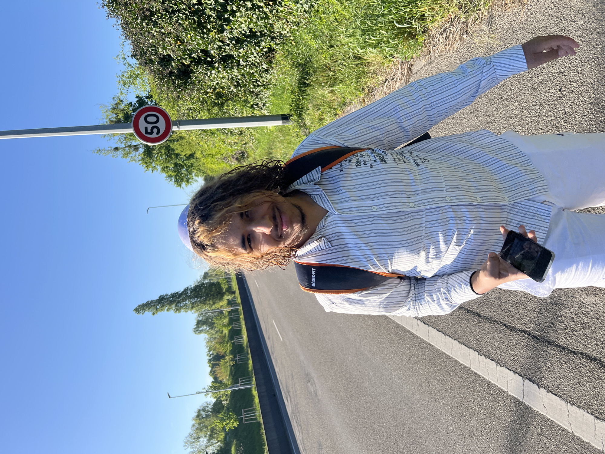 A person is standing on a road in Hoenheim, France, holding a smartphone in their right hand. They are wearing a vertically striped button-up shirt, white pants, and a blue head covering. A black backpack strap is visible on their shoulder. A "50" speed limit sign is to the left of the person. The road stretches into the distance, bordered by trees and streetlights. The sky is clear and blue.