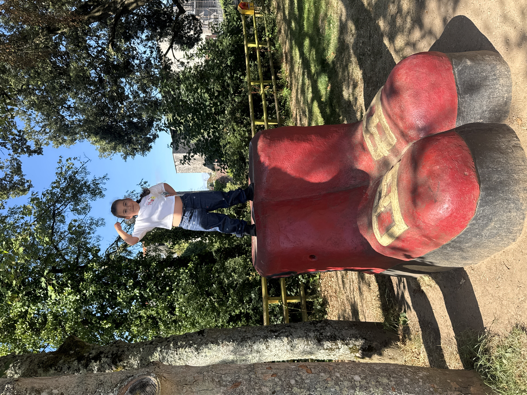 A young girl is standing on a large, red, novelty boot sculpture in a park. She is wearing a white t-shirt and blue jeans, and she is flexing her bicep. The boot sculpture is red with yellow accents and appears to be made of concrete. The park has trees, grass, and a yellow fence in the background. In the distance, there are buildings, possibly part of Cahir Castle. The weather is sunny with a clear blue sky.