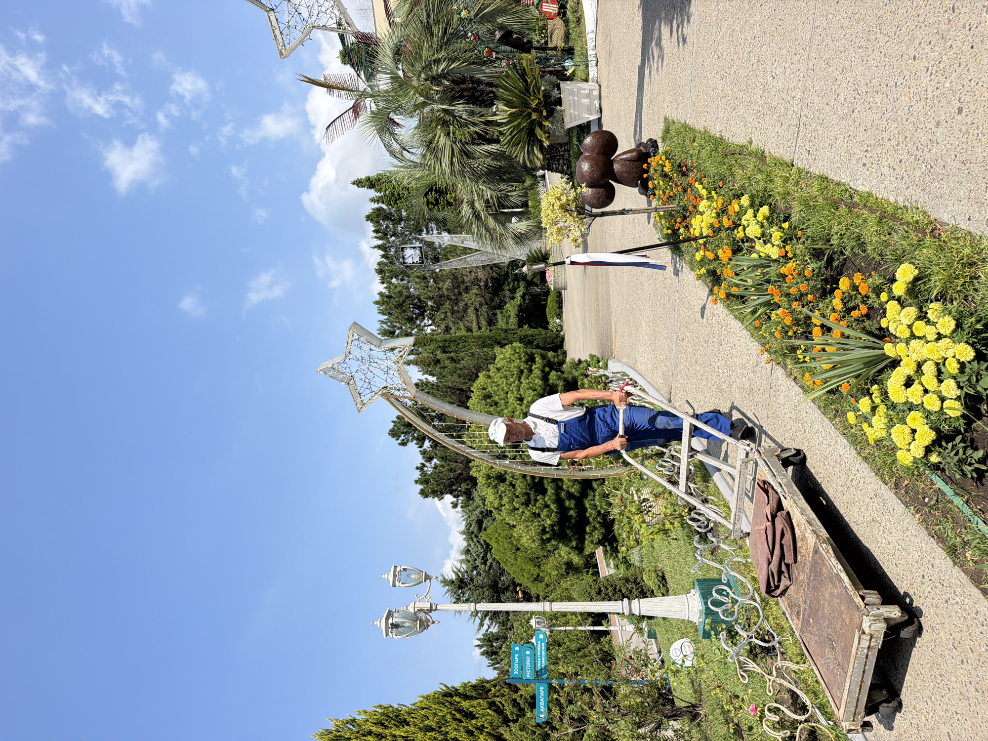 A man wearing blue overalls and a white cap pushes a flatbed trolley along a paved pathway lined with flowerbeds. The man is positioned in the center of the frame, moving from right to left. The trolley, which appears old and weathered, carries a folded brown cloth. To the left of the pathway, there is a lamppost with two lamps and a signpost with several directional signs. In the background, various trees and bushes create a lush, green environment. Large decorative star-shaped structures, likely illuminated, are visible, suggesting a festive or park-like setting. A small, dark brown, rounded statue, possibly depicting an animal, is situated in one of the flowerbeds. The sky is clear blue with scattered white clouds. The scene appears to be outdoors, possibly in a public park or recreational area within Sochi, Russia, given the presence of palm trees and the overall subtropical atmosphere.