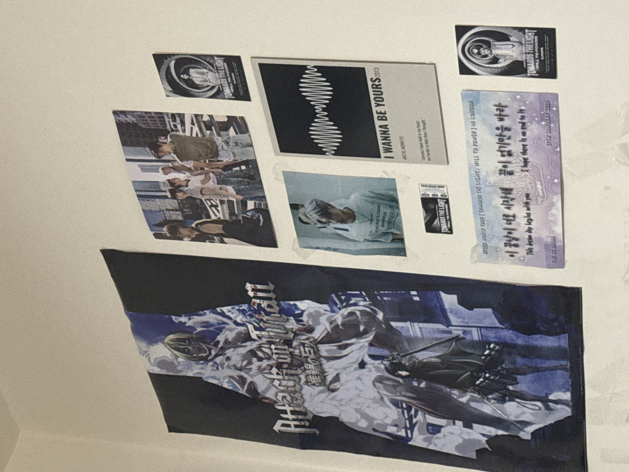The image shows a collection of posters and memorabilia adhered to a light-colored wall.
Items include:
* A large black and white poster featuring a stylized graphic with text in a language other than English.
* Two smaller "TOWARDS THE LIGHT" posters with the words "WILL TO POWER" and "EUROPE."
* A poster for the Arctic Monkeys' song "I WANNA BE YOURS" (2013).
* A group of people in an urban setting.
* A ticket stub or print with Korean text and the date "27TH JANUARY 2025."
The precise location within Sofia, Bulgaria, is not identifiable from the image.