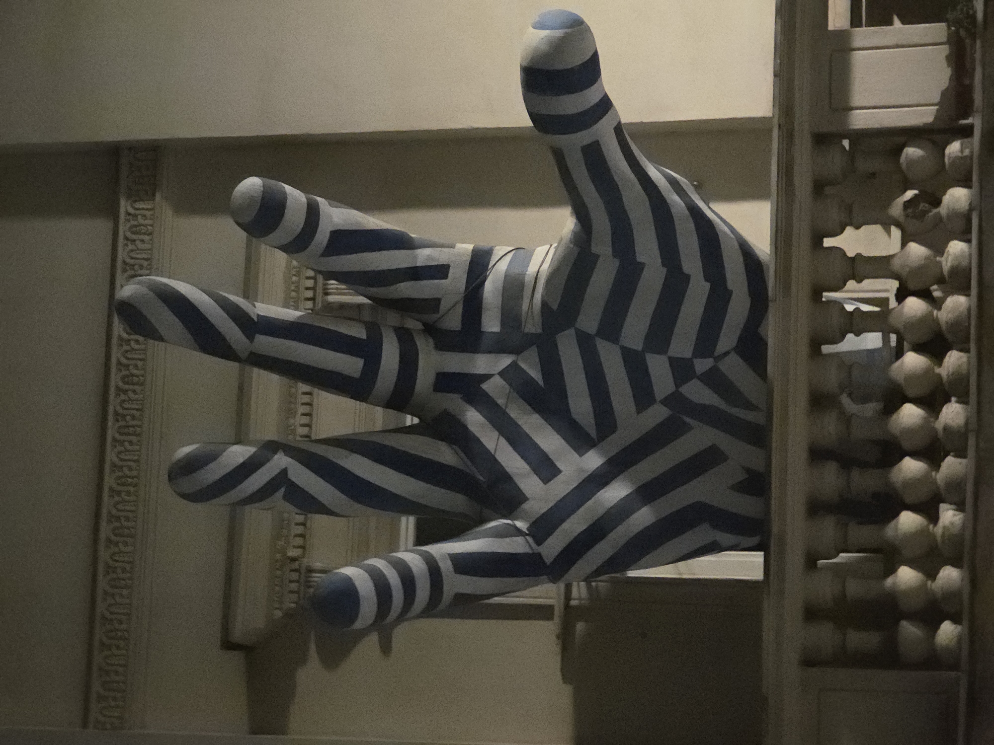 The image shows a large, striped blue and white hand sculpture reaching out of a building. The sculpture is positioned next to a doorway with a lattice-like decorative element on the right side. The building's facade has a classical architectural style, including decorative moldings. The scene is dimly lit. The image depicts the artwork "Palm Tree" (Pozdrowienie z Alej Jerozolimskich) located at Aleje Jerozolimskie in Warsaw, Poland.