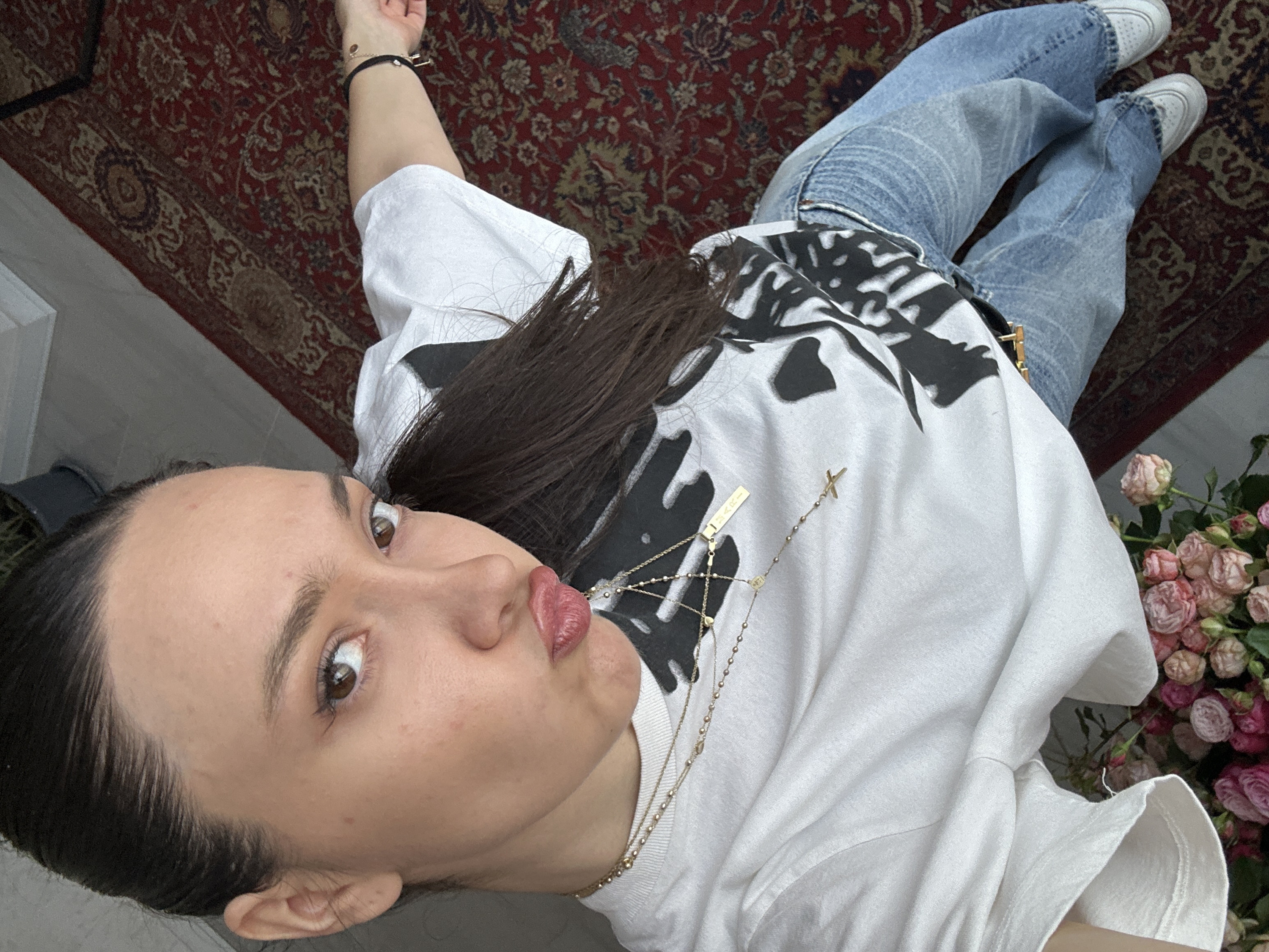 A young adult female is depicted in a full-body selfie taken indoors within Yerevan, Armenia. She has dark hair pulled back and brown eyes, looking directly at the camera with a "duck face" expression. Minor blemishes are visible on her forehead and left cheek. She wears a white short-sleeved t-shirt featuring a black graphic design, light blue denim jeans, and white low-top athletic shoes. Her accessories include multiple gold-toned necklaces, one of which is a rosary-style chain with a small cross pendant and a rectangular pendant inscribed "MARI," and a black wristband with charms on her left wrist. In the lower-left foreground, a bouquet of pink and peach miniature roses is present. The flooring consists of an ornate red rug patterned with cream and beige designs. A light grey wall or floor transition and a dark potted plant are visible in the background. The scene appears to be a residential or private interior.
