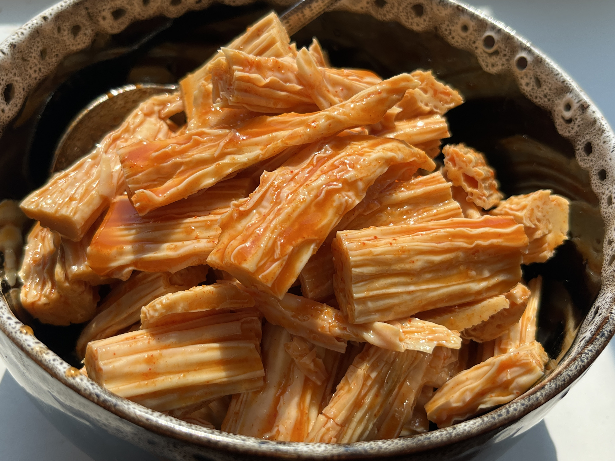 A close-up, eye-level shot shows a bowl filled with a spicy, orange-marinated food. The food appears to be cut into short, cylindrical pieces with a textured, layered surface, possibly a type of dried tofu or a similar ingredient. The orange marinade coats the food, with some glistening oil visible. The bowl has a rustic, speckled brown and white rim. The background is blurred, suggesting an outdoor or well-lit environment. No people or specific location details are discernible.