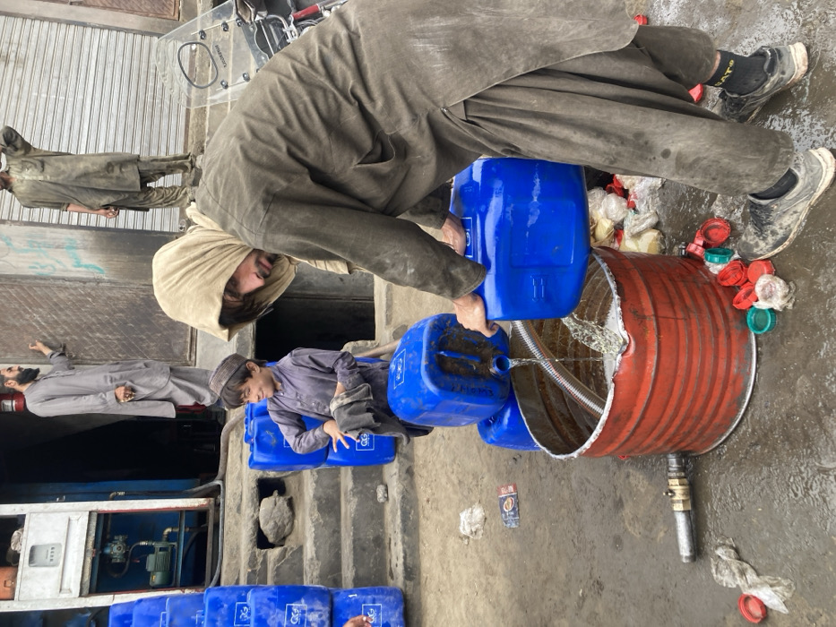 This image depicts an outdoor scene in Quetta City Tehsil, Pakistan, showing a liquid transfer operation amidst an urban environment.
In the foreground, a man, likely in his 20s, wearing a dirty, light brown shalwar kameez and a beige head covering, is bent over, pouring a clear liquid from a blue plastic jerrycan into a large, open, red metal barrel positioned on the wet, muddy ground. The liquid is actively splashing into the barrel, which contains a flexible hose and has a metal spigot attached to its lower side. The man's footwear consists of dark socks and light-colored, dirty sneakers. Scattered on the ground around the barrel are numerous red, green, and white plastic bottle caps, plastic bags, and a discarded wrapper.
To the left, slightly behind the pouring man, a young boy, approximately 7-10 years old, wearing a dirty, dark purple shalwar kameez and a dark cap, is seated on a stack of blue plastic jerrycans. He is holding a dark cloth in his hands and looking towards the pouring activity with a slight smile. More blue jerrycans, some bearing a white "GG" logo, are stacked vertically near the boy and extend out of the frame to the left.
In the elevated background, two adult men are visible. On the left, a man with a dark beard, wearing a light gray shalwar kameez, stands on a raised concrete step or platform near an open doorway. A white fire extinguisher is mounted on the wall behind him. Adjacent to the doorway, an open niche reveals what appears to be a water pump or meter. Further to the right, another man, dressed in a dirty, light brown shalwar kameez, stands on a similar elevated surface behind a partially lowered metal shutter, observing the scene. A portion of a motorcycle windscreen is visible on the far right. The overall ground is uneven, damp, and dirty.