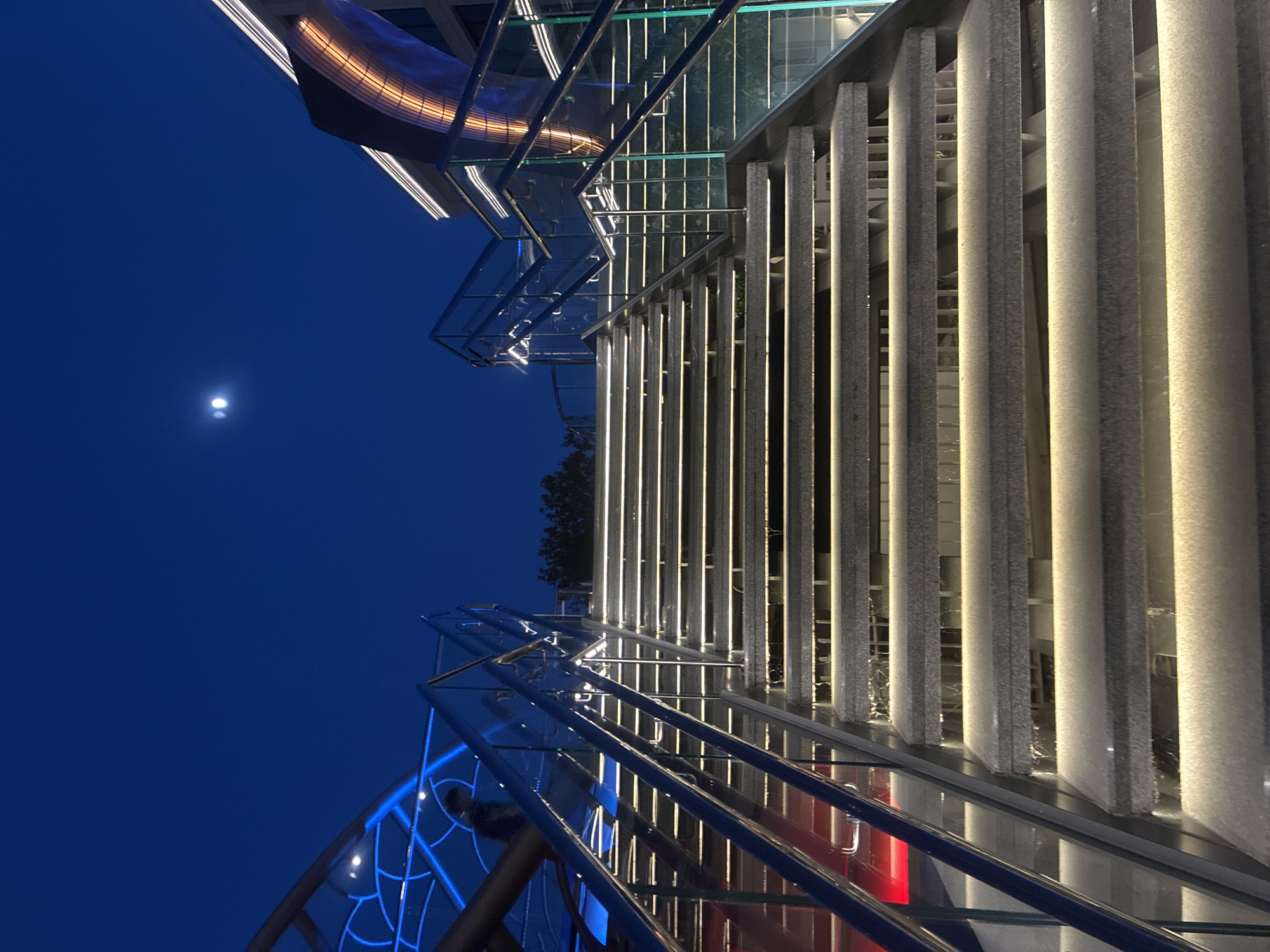 The image depicts an exterior view of a modern building at night, showcasing its architectural design and lighting. The primary subjects are the building's facade, featuring a series of vertical, textured stone elements illuminated by warm, vertical lights, and a glass structure with metal railings. The scene is set against a deep blue night sky, with a full moon visible in the upper left quadrant of the image.
The building appears to be a contemporary structure, possibly a hotel or a commercial space, given its sleek lines and sophisticated lighting. The vertical elements on the facade create a sense of rhythm and depth, while the glass and metal components add a touch of modern elegance. The bright blue geometric patterns within the glass structure on the lower left suggest decorative lighting or stained glass. A hint of red is visible behind the glass railing in the lower right, possibly indicating interior lighting or decorative elements.
The time of day is clearly night, indicated by the dark sky and the prominent artificial lighting. The weather appears clear, with the moon visible and unobscured. While the specific location within Sochi, Russia, cannot be precisely identified from the image alone, the architectural style suggests a developed urban or resort area. There is no specific activity or event taking place, and the image focuses on the static architectural elements. No text is visible in the image. The overall mood conveyed is one of sophisticated urban ambiance and modern architectural beauty illuminated by the night.