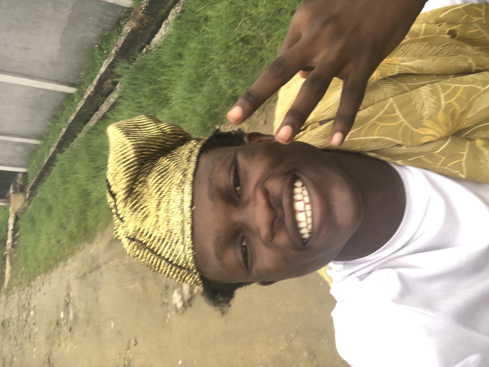 A medium shot captures a young Black man smiling and holding up two fingers in a peace sign. He is wearing a gold, textured cap and a white t-shirt, with a gold patterned cloth draped over his shoulder. The background is blurred, showing a dirt path, green grass, and a grey wall. The scene appears to be outdoors, possibly in Ado, Nigeria, given the context.