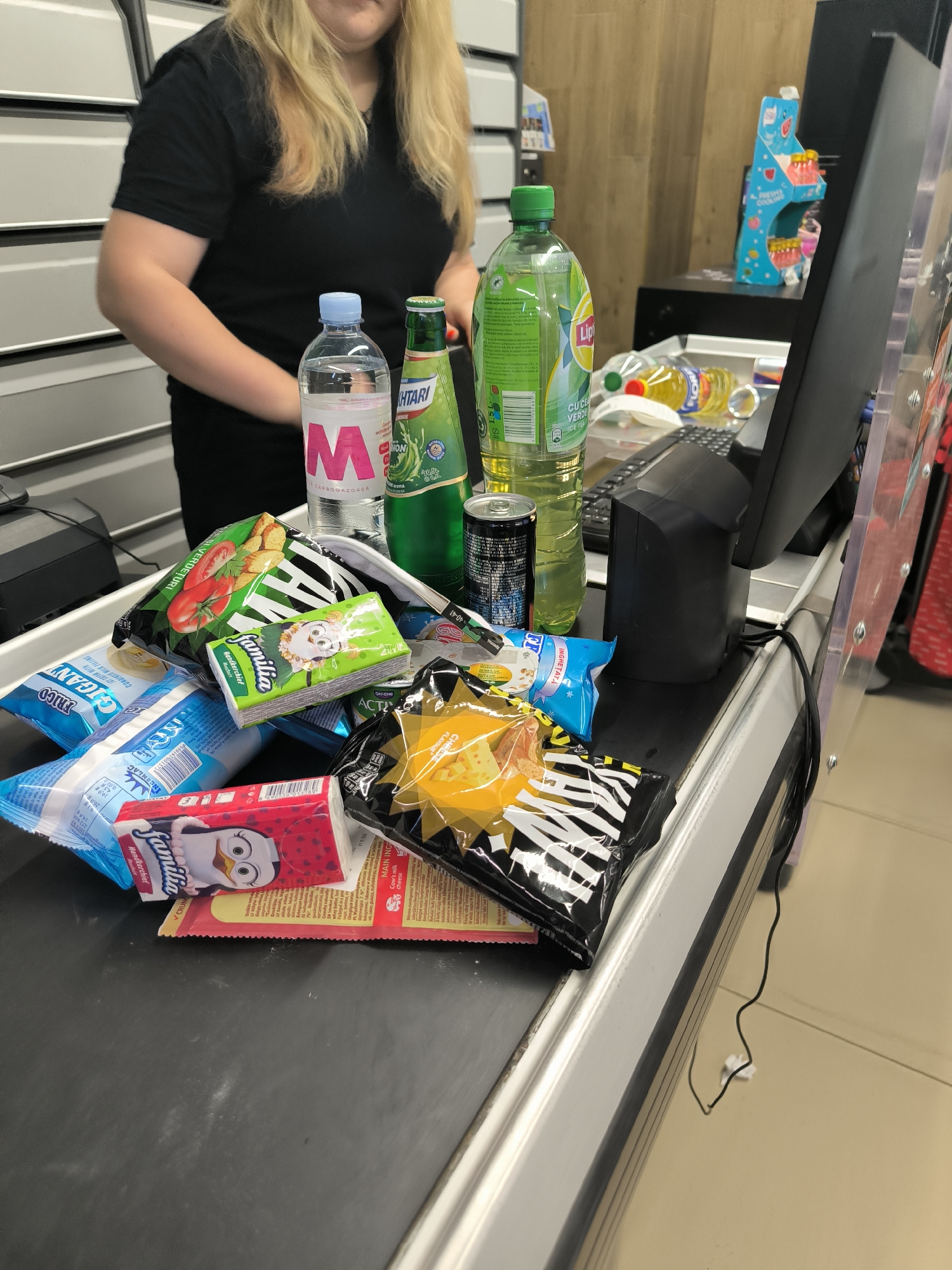The image captures a close-up, eye-level view of a supermarket checkout counter in Chișinău, Moldova. A cashier, a woman with blonde hair wearing a black t-shirt, is partially visible in the background, her hands out of frame. The focus is on the items placed on the conveyor belt, which are a mix of snacks and beverages. These include: two bottles of Lipton Ice Tea (one green, one with a yellow liquid), a bottle of "M" branded water, a can of soda, a bag of chips with a tomato design, a small packet of "Familia" brand handkerchieves with a cartoon bird, another packet of "Familia" handkerchieves with a cartoon owl, a blue bag of what appears to be wet wipes, and a bright yellow bag of snacks with a black and white striped pattern. The scene is set in a brightly lit supermarket, evidenced by the uniform grey shelving in the background and the polished surface of the checkout counter. The presence of a computer monitor and a payment terminal suggests an active point of sale.