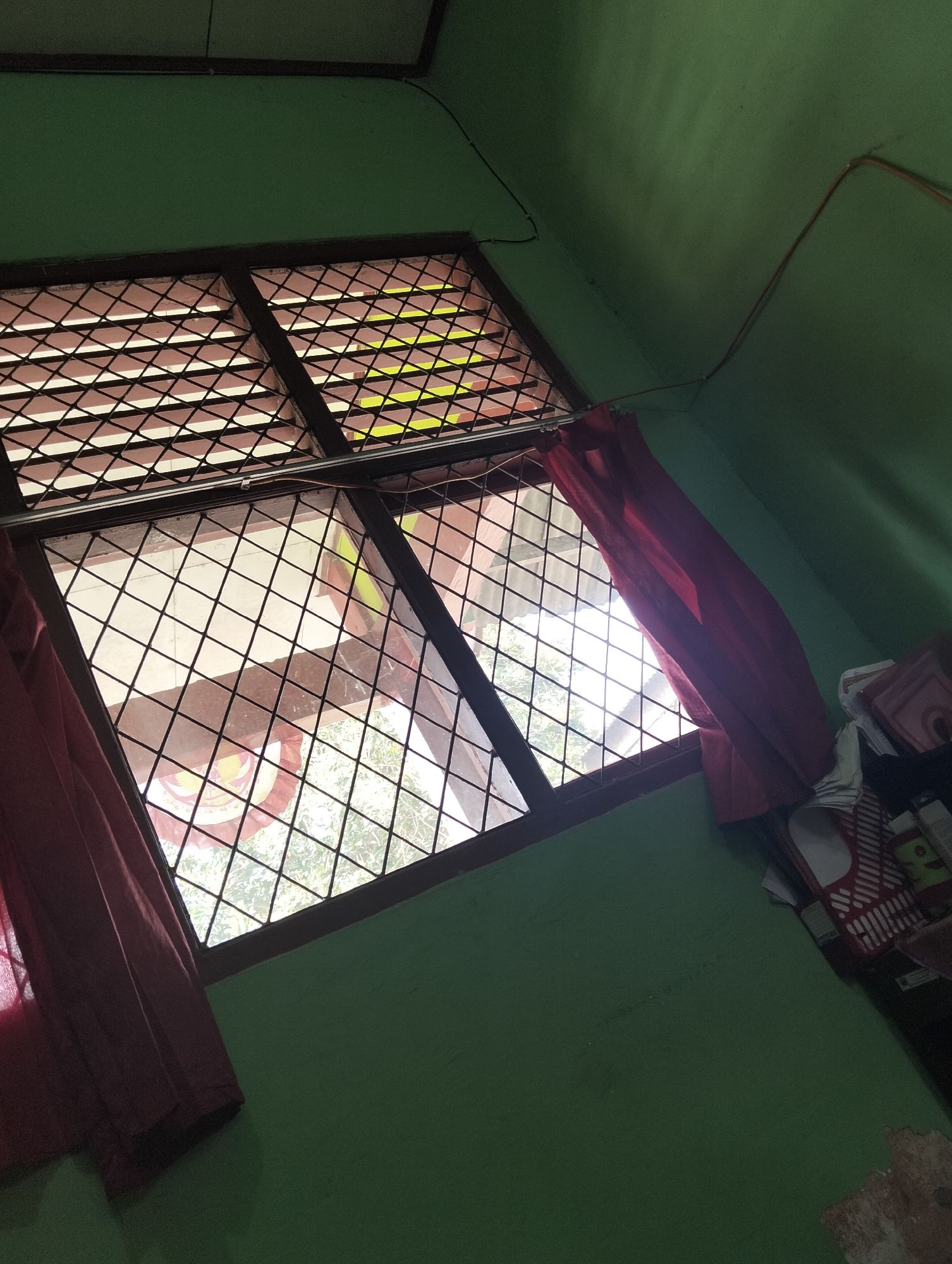 The image depicts an interior view of a room with green walls, angled to capture a window. The window features a diamond-patterned metal grate. Red curtains hang on either side of the window, partially obscuring the view outside, which appears to be trees and a corrugated roof. To the right of the window, a shelf is visible with several items, including what looks like a red and white striped basket or container. Electrical wires are visible running along the ceiling and down the wall. The lighting suggests it is daytime, with sunlight streaming through the window. The room appears to be in a residential setting, possibly in Banjar Agung, Indonesia, based on the prompt's context, though this cannot be definitively confirmed from the image alone. There are no people or animals visible in the image.