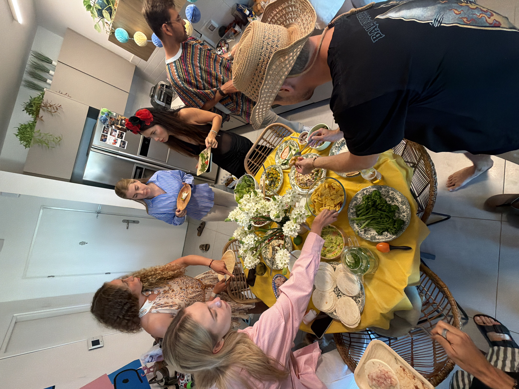 An overhead shot captures a gathering of at least six people around a circular table laden with food and drinks, possibly in a kitchen or dining area within a Tel Aviv apartment. The table is draped with a bright yellow cloth, on which various dishes are arranged, including what appears to be guacamole, corn chips, tortillas, and a plate of cooked green vegetables. A centerpiece of white flowers adds a decorative touch.
The individuals present exhibit a range of attire and engagement with the meal. Two women, one with long blonde hair wearing a pink striped shirt, and another with curly brown hair in a patterned dress, are actively interacting with the food. A third woman, dressed in a blue striped shirt, holds a small plate. A man wearing a colorful, patterned shirt stands near a refrigerator adorned with photographs. Another man, positioned in the foreground and seen from the back, wears a black t-shirt with a graphic design and a straw hat. The environment suggests a casual, communal meal, possibly a brunch or lunch, given the bright lighting and relaxed atmosphere. The presence of a refrigerator with photos implies a residential setting, likely a home in Tel Aviv.