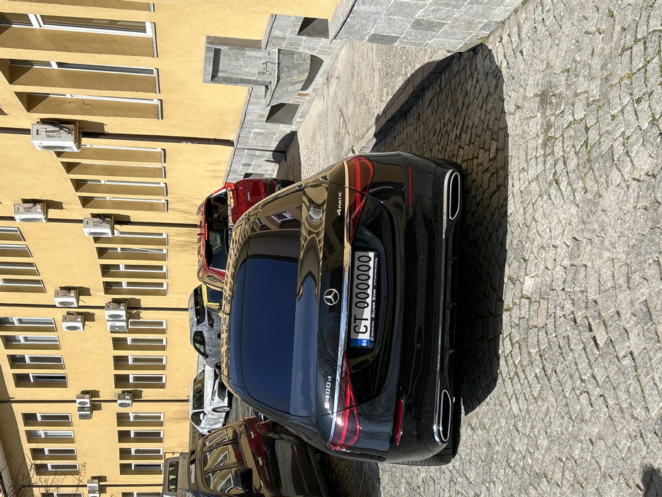 The scene depicts a parked black Mercedes-Benz S400d 4MATIC sedan, viewed from the rear, on a cobblestone surface in Plovdiv, Bulgaria. The vehicle features tinted rear glass, chrome trim, and dual integrated exhaust tips. Its Bulgarian license plate reads "BG CT 000000", framed by a "Silver Star" dealership plate holder.
Behind the Mercedes, several other vehicles are parked in a row, partially obscured. These include a red SUV, a white sedan, and at least one other grey sedan. To the left, a dark-colored SUV or station wagon is partially visible.
To the right of the Mercedes, a multi-story building with a bright yellow facade is present. The building features numerous vertical rectangular windows with white frames arranged in two rows. Multiple external air conditioning units are mounted below these windows, with visible conduits. A low wall, clad in grey stone, runs along the base of the building. This wall incorporates a stone water fixture with a basin and spigot, as well as recessed sections, possibly planters or benches, also clad in stone.
The environment is illuminated by direct sunlight, casting distinct shadows on the cobblestones and the building's facade. No individuals are visible.