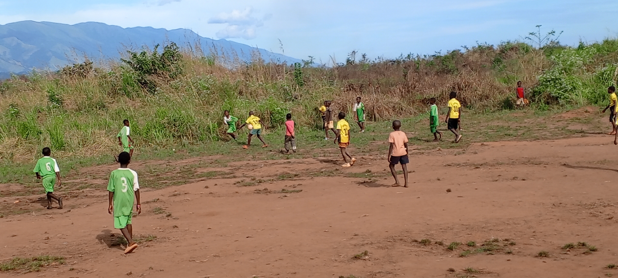 In Ngalula, Democratic Republic of the Congo, a group of boys is engaged in a soccer game on a dirt field. Some of the boys are wearing green jerseys with numbers on the back, while others are in yellow jerseys, also numbered. One boy is wearing an orange t-shirt and navy shorts. The playing surface is bare earth, transitioning into tall grasses and scrub brush at the edges of the field. In the background, there are distant, blue mountains under a light blue sky with scattered clouds.