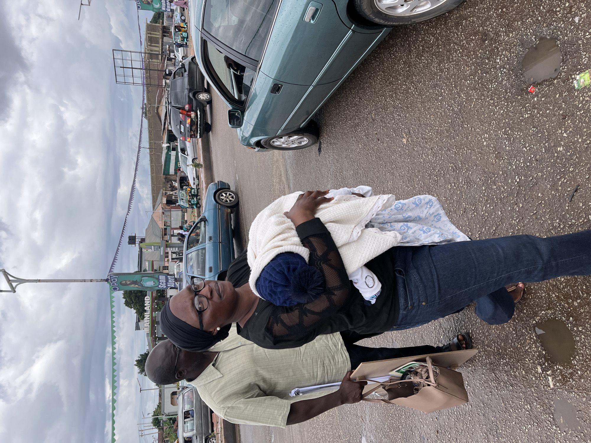 In Gura Top, Nigeria, a man wearing a light-colored short-sleeved collared shirt and dark pants stands to the left of a woman. The woman, dressed in a black top with sheer sleeves and blue jeans, wears glasses and a black headscarf. She is holding a bundled infant, covered in a white blanket with blue patterns and a blue knitted hat. The man carries a brown tote bag and a large, flat board. They are on a wet asphalt road, with visible puddles. Behind them, a teal sedan is parked, and other vehicles, including a black minivan and a blue sedan, are present on the road. Several buildings and a tall lamppost with signage are visible in the background under a cloudy sky.