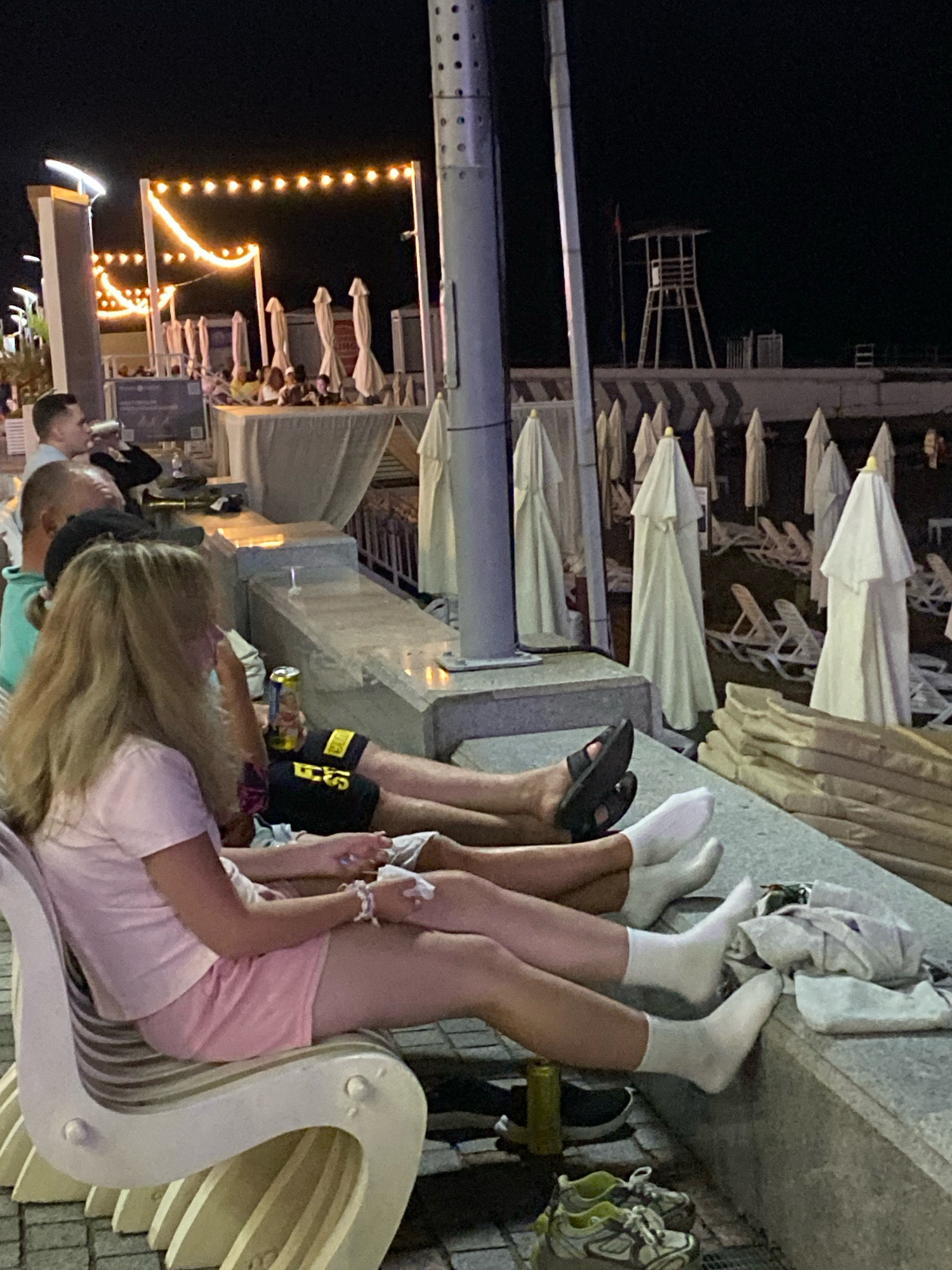 A group of people are seated on a bench and on the ground in an outdoor setting at night, possibly a cafe or promenade near a beach. String lights are visible in the background, casting a warm glow. Beach umbrellas and lounge chairs are stacked in the background, suggesting the location is a beachside resort. One person in the foreground is wearing a pink t-shirt and pink shorts, with their legs crossed. Another person is wearing black shorts with yellow lettering and black sandals. A third person is wearing white socks. There is a beer can on the ground near the people. In the far background, a lifeguard tower is visible. The overall scene depicts a relaxed evening gathering in a tourist area, likely in Sochi, Russia, given the presence of beach amenities and the general atmosphere.