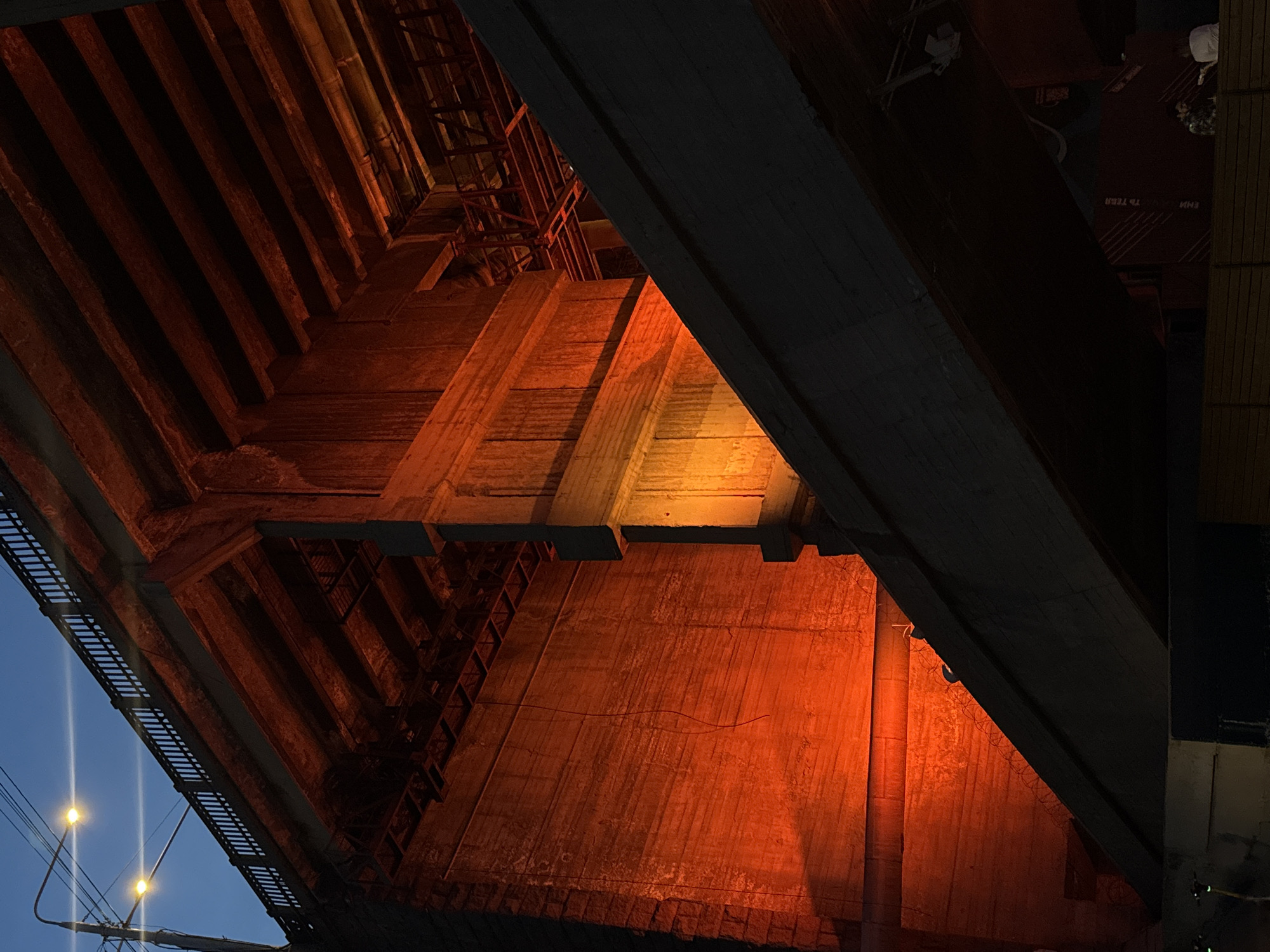 The image shows the underside of a concrete bridge illuminated with warm orange and yellow lights, creating a stark contrast against the twilight sky. The bridge structure is robust, with visible concrete supports and beams, some of which are weathered and show signs of age. A dark, angled structure, possibly another part of the bridge or an elevated roadway, cuts across the frame. The lighting casts dramatic shadows, highlighting the textures and forms of the concrete. In the distance, under the bridge, there appear to be some figures and structures, suggesting an urban environment. The overall impression is one of industrial architecture bathed in artificial light, with a sense of depth and scale. The specific location in Krasnoyarsk, Russia, is not identifiable from the visual information alone.