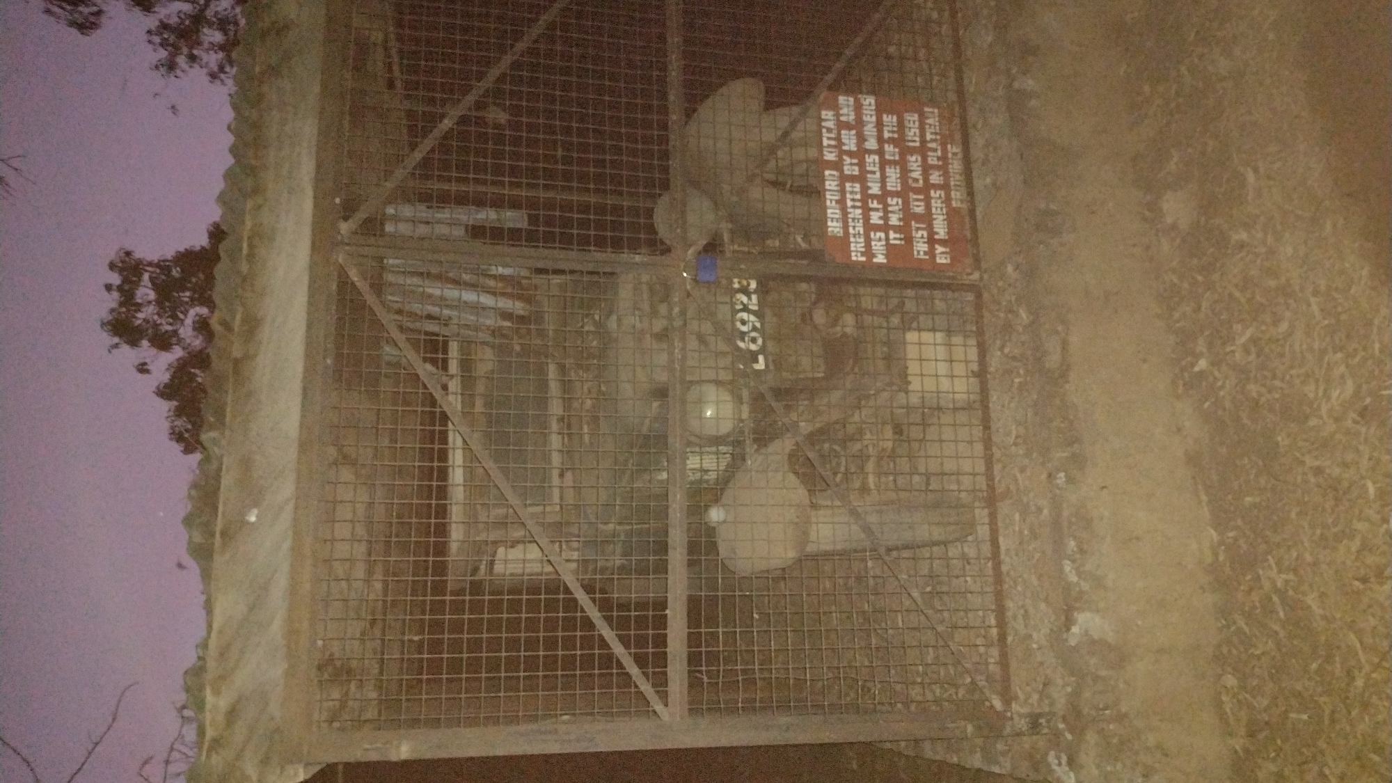Captured at night, the image reveals a Bedford Kitcar encased within a protective cage. The Kitcar, designated with the number "26923", is accompanied by an adjacent sign. The sign indicates that the vehicle was presented by Mr. and Mrs. M.F. Miles, described as miners. It further states that the Bedford Kitcar was among the initial kit cars used by miners in Plateau Province, suggesting that the vehicle is located in Jos, Nigeria.