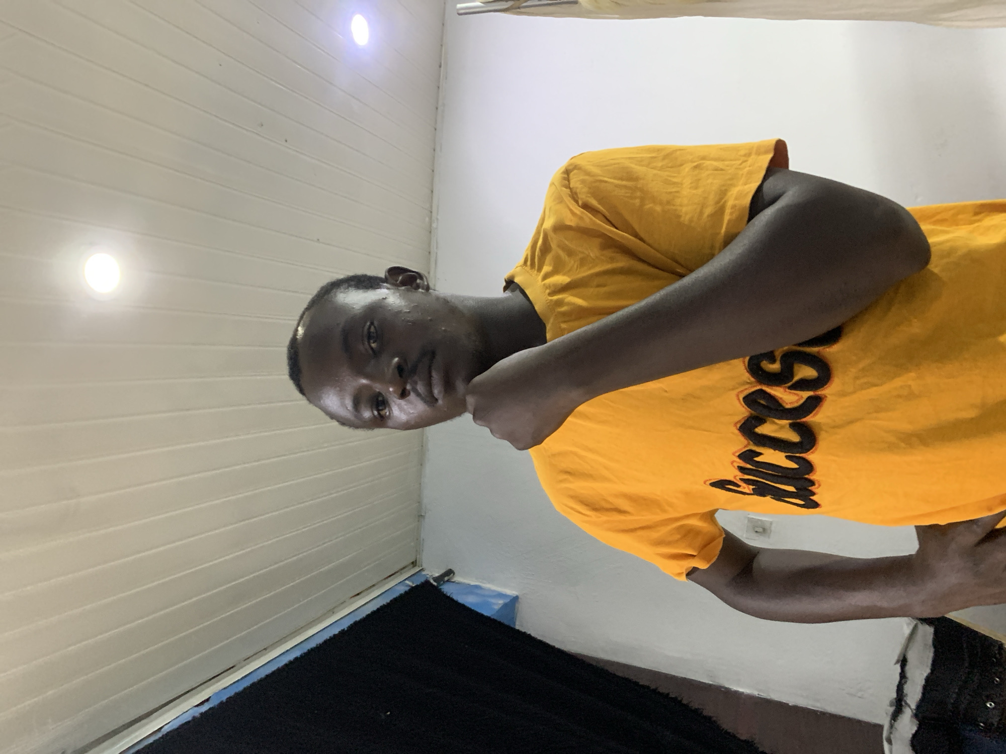 A young man with dark skin and short dark hair is depicted indoors in a vertically oriented image. He wears a bright yellow t-shirt with the word "Success" printed in black, stylized font across the chest. He looks directly at the camera with a neutral expression, his right arm bent with his hand positioned near his face.
The background features a room with distinctive elements. To the man's left is a section of white or cream-colored horizontal paneling, possibly a ceiling or upper wall, fitted with two bright, circular light fixtures that emit a strong white glow. To his right, a plain white wall is visible. In the lower left corner, a textured, dark-colored material, resembling a rug or curtain, rests against a blue-painted baseboard or trim. The lighting is entirely artificial, suggesting an indoor setting. This casual portrait was taken in Jos, Nigeria.