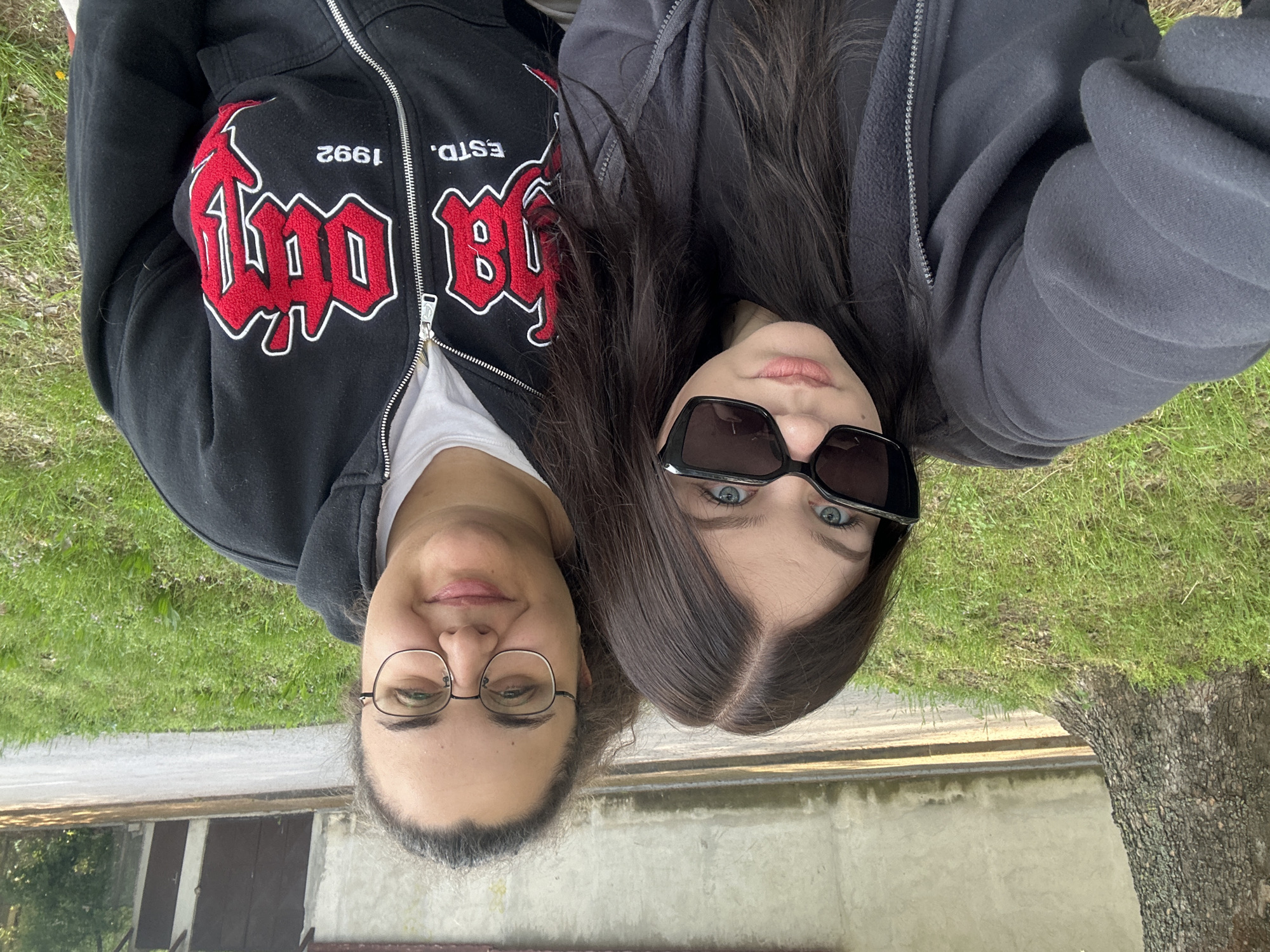 Two women are posing close together for a selfie-style photograph, with the camera held in a somewhat upside-down orientation. The woman on the left is wearing glasses and a black zip-up hoodie with red and white lettering. The woman on the right has dark hair and is wearing large black sunglasses and a gray zip-up hoodie. They are positioned in front of a grassy area, with a concrete structure and a tree visible in the background. The scene appears to be outdoors. While I do not have the information to be certain, based on visual features of the vegetation, buildings, and the description provided, the location may be in or around the city of Beloslav, Bulgaria.