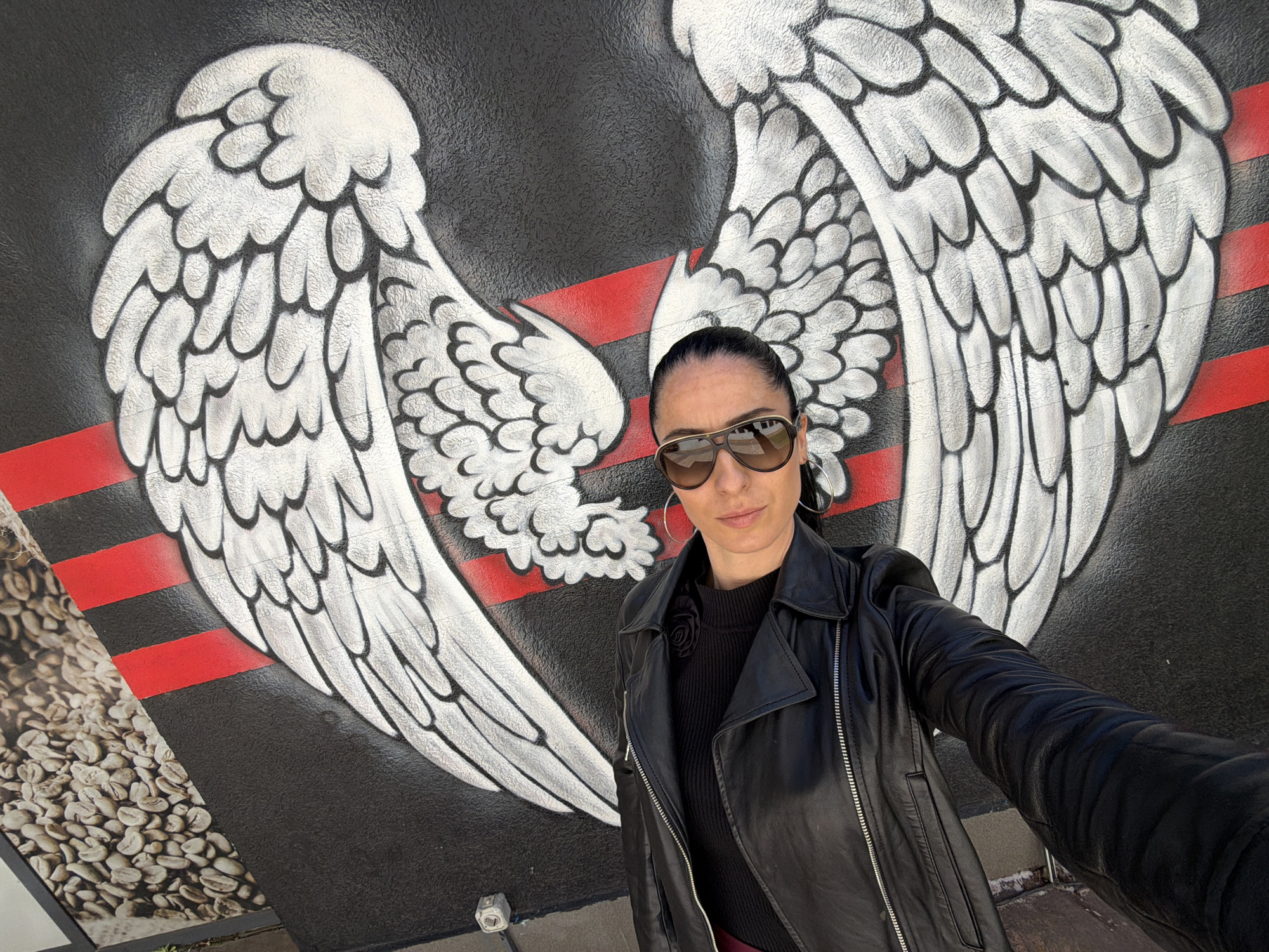 A woman is posing for a selfie or photograph in front of a striking wall mural in Pleven, Bulgaria.
The central subject is a woman with dark hair, pulled back into a ponytail. She is wearing large aviator-style sunglasses that cover her eyes, large silver hoop earrings, a black leather motorcycle jacket, and a black ribbed top underneath, which features a black rose-like embellishment on the left side of her chest. Her expression appears neutral and composed. She is holding the camera herself, with her right arm extended, visible in the bottom right corner of the frame.
The setting is an outdoor location, likely a street or café exterior, featuring a large, distinctive mural on a wall. The mural depicts a pair of large, stylized white wings with prominent black outlines and feather details, set against a black background. Two horizontal red stripes run across the black background and under the wings. To the left side of the mural, there is an additional section with an image of raw coffee beans. The wall surface appears to have a slightly textured finish.
The activity is clearly a photo opportunity, with the woman positioning herself to appear as if the wings are behind her. The bright, natural lighting suggests it is daytime. There is no visible text in the image.