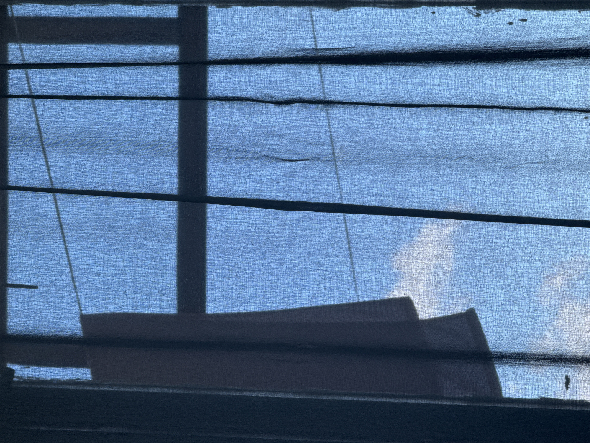 The image provides a close-up, backlit view of a window covered by a translucent fabric blind, set in Chaikovsky, Russia. The blind is a light blue-grey color, displaying a visible woven texture and distinct horizontal pleats or folds. Strong daylight from outside illuminates the fabric, making it somewhat transparent and revealing shadows cast upon it.
Dark, defined silhouettes of parts of a window frame, including vertical and horizontal bars, are prominent on the blind. In the lower section of the image, silhouettes of objects positioned on a windowsill or outside ledge are visible. These objects appear angular and indistinct, resembling architectural elements or containers. Through the upper right portion of the translucent fabric, faint lighter areas suggest the presence of clouds or a bright sky beyond the window. Thin, light-colored cords, likely part of the blind's operating mechanism, are visible on the left side. The scene focuses on the interplay of light and shadow on the textured fabric, creating a serene, observational mood. No people, activity, or visible text are present.