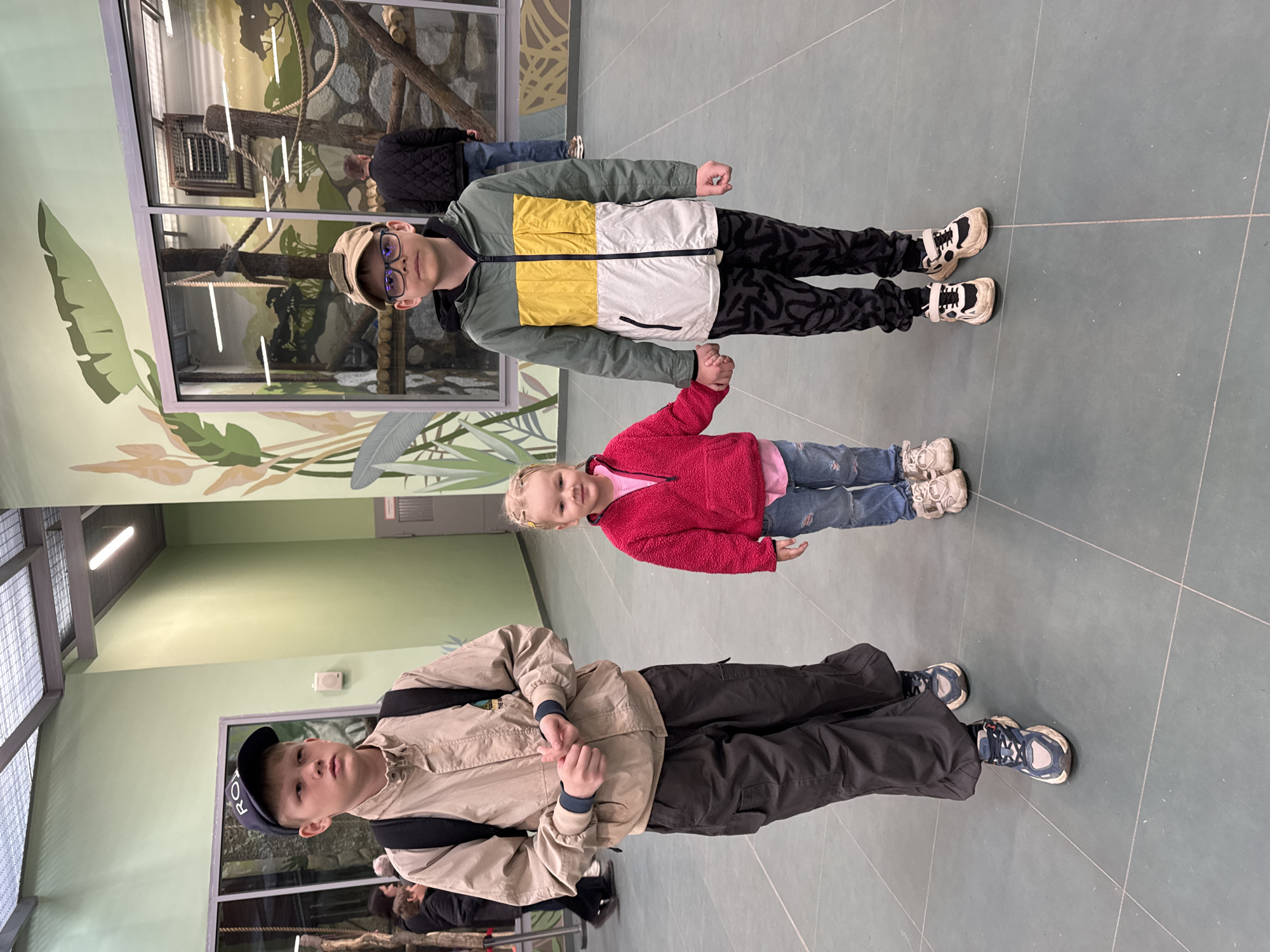 Three children are standing in an indoor space with a light green floor and walls, likely a zoo or wildlife exhibit based on the painted murals of tropical plants and animal enclosures visible in the background. At the bottom of the image, a boy with a dark baseball cap and a tan jacket is wearing a black backpack. In the middle, a young girl in a pink fleece jacket and jeans is holding hands with the boy at the top of the image. The boy at the top is wearing a yellow, white, and green jacket, patterned pants, and glasses. The location is likely in the city of Perm, Russia, considering all elements.
