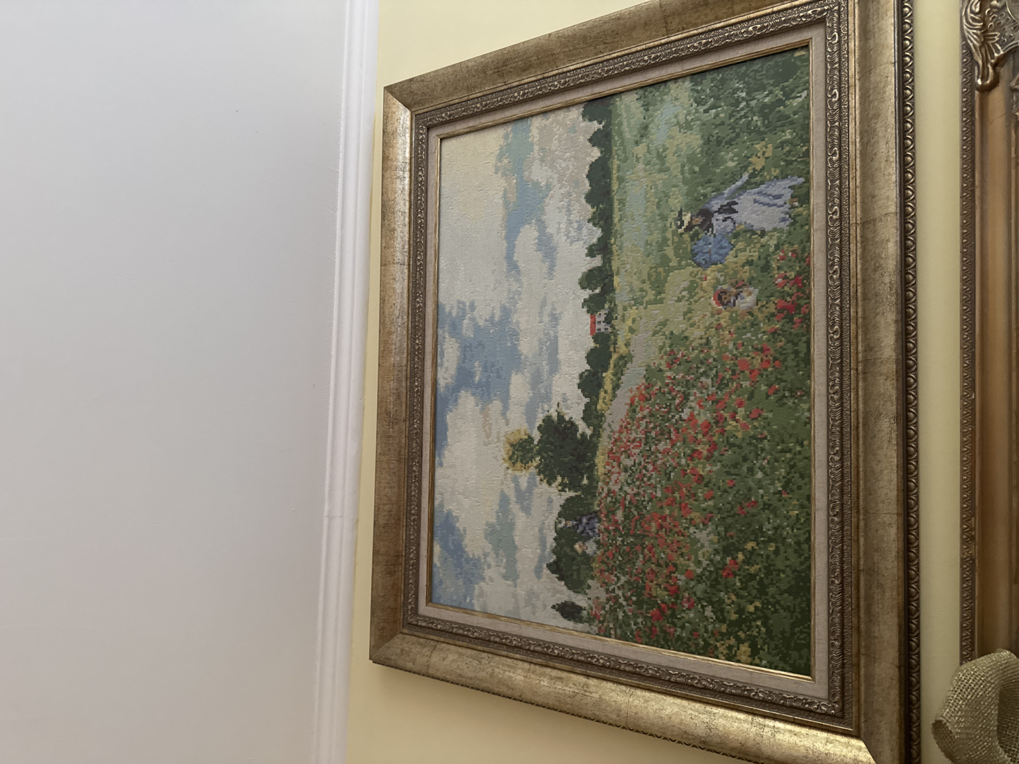 The media file displays a framed piece of textile art, possibly a tapestry or needlepoint, hanging on a light yellow wall. The artwork depicts a vibrant, idyllic landscape scene. In the foreground, a vast field of lush green grass is dotted with numerous bright red flowers, reminiscent of poppies. Further back, trees and foliage create a natural boundary, and a small, red-roofed house is visible in the distance. The sky above is a mix of light blue with soft white clouds.
Two human figures are visible within the artwork: a woman in a blue dress, wearing a hat, appears to be standing or walking in the field on the right side of the painting, and a smaller figure, possibly a child, is seen near her. The overall impression is a peaceful, pastoral setting.
The artwork is encased in an elaborate, ornate gold-colored frame with intricate decorative patterns. To the left of the framed artwork, a white door frame or trim is visible against a plain white wall. To the right, part of another gold-framed object is seen, suggesting other decor on the wall. A piece of burlap or similar textured fabric is partially visible in the bottom right corner, near the other frame. The room appears well-lit, highlighting the metallic sheen of the frames and the vibrant colors of the artwork. No visible text is present in the image.