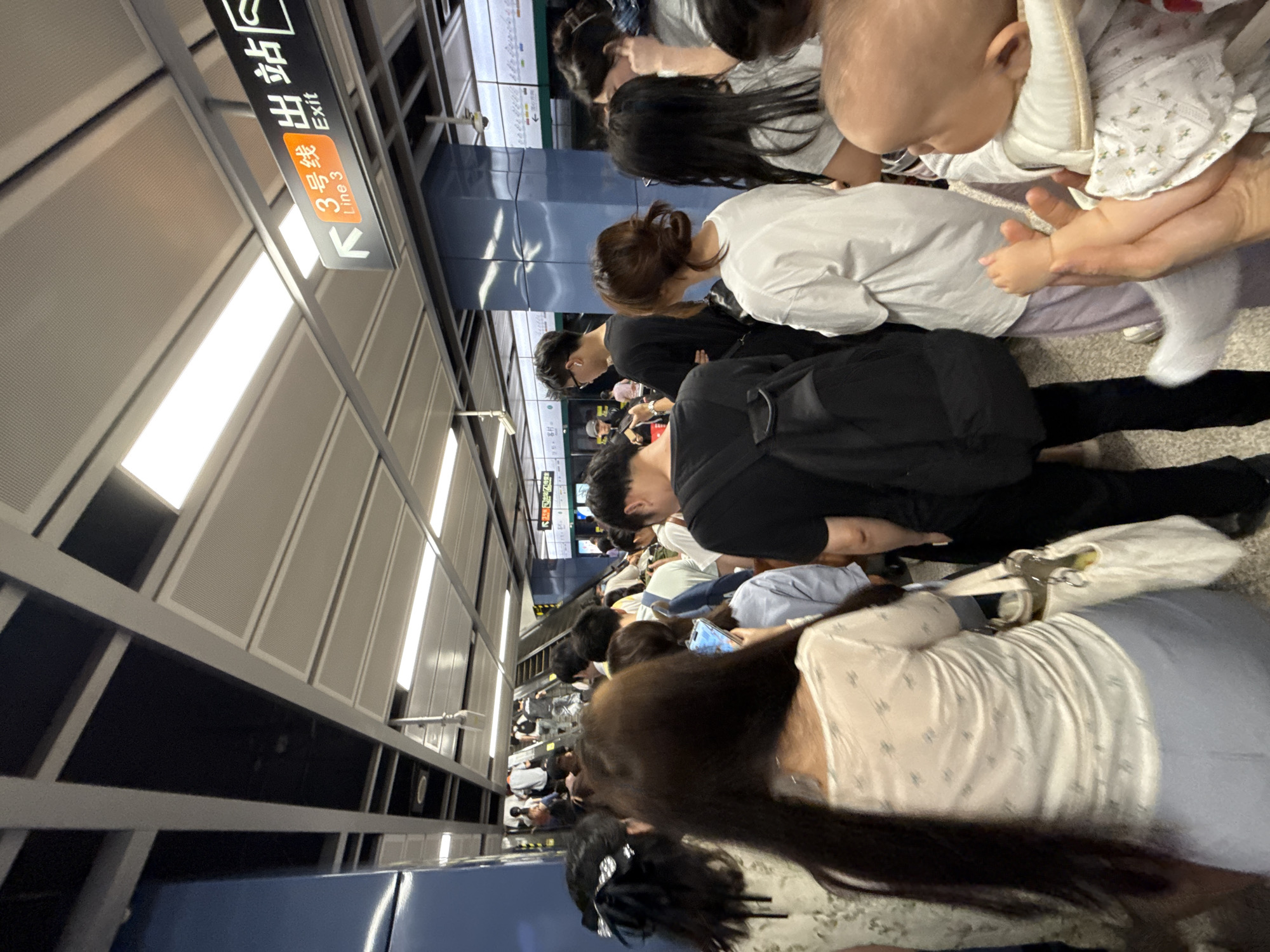 A crowd of people is seen on a subway platform in Haizhu District, China, with a sign overhead indicating "Line 3" and "Exit" with an arrow pointing to the left. The platform is well-lit by fluorescent ceiling lights. In the foreground, a woman with long dark hair is visible from behind, wearing a patterned white blouse and a light blue skirt, carrying a white tote bag. To her right, a baby is being held, facing away from the camera, with its legs and feet exposed. Other individuals are standing and walking, some facing escalators in the background. The overall scene suggests a busy public transportation hub during peak hours.