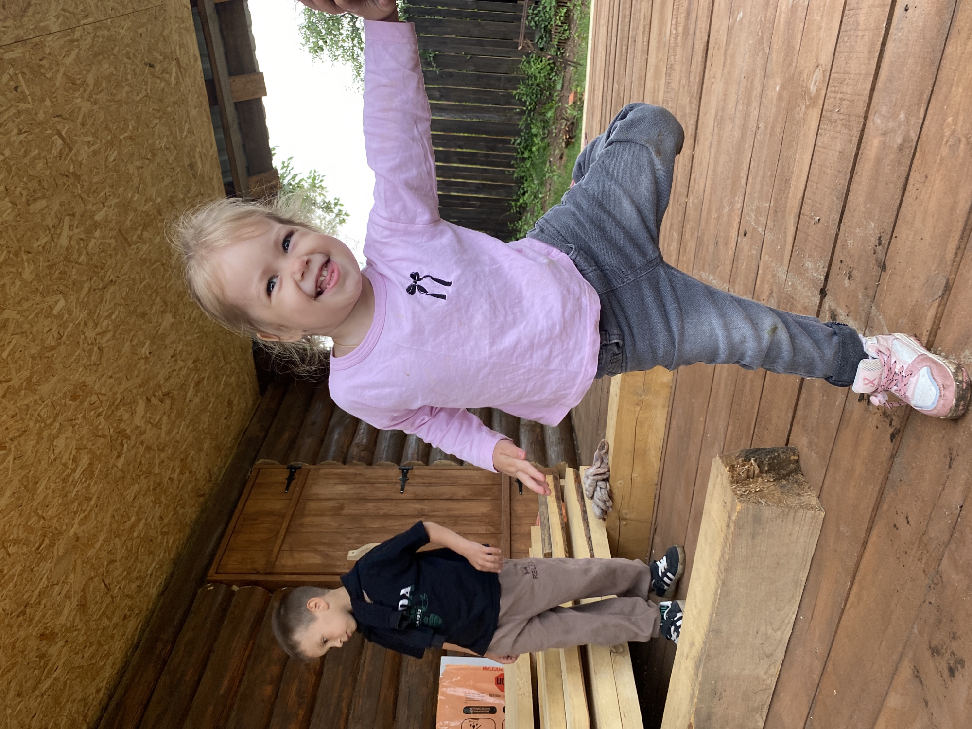 In this vertically oriented outdoor photo, two young children are the subjects on a wooden deck. The girl in the foreground, with blonde hair, is wearing a pink long-sleeved shirt and grey jeans. She is smiling widely and appears to be reaching out. Her pink and white sneakers are visible. Behind her and lower in the frame, a boy with short brown hair is wearing a black t-shirt and brown sweatpants. He is looking down, and his black and white sneakers are visible. They are on a wooden deck, with wooden walls and a door visible in the background. The setting appears to be rustic, possibly a porch or a wooden structure in a yard. The lighting suggests it is daytime, and the weather appears to be mild. The scene is casual and playful, with the girl's expression conveying joy. A visible piece of wood is in the foreground, and some gardening equipment or supplies are also visible near the boy. The location is Chaikovsky, Russia.
